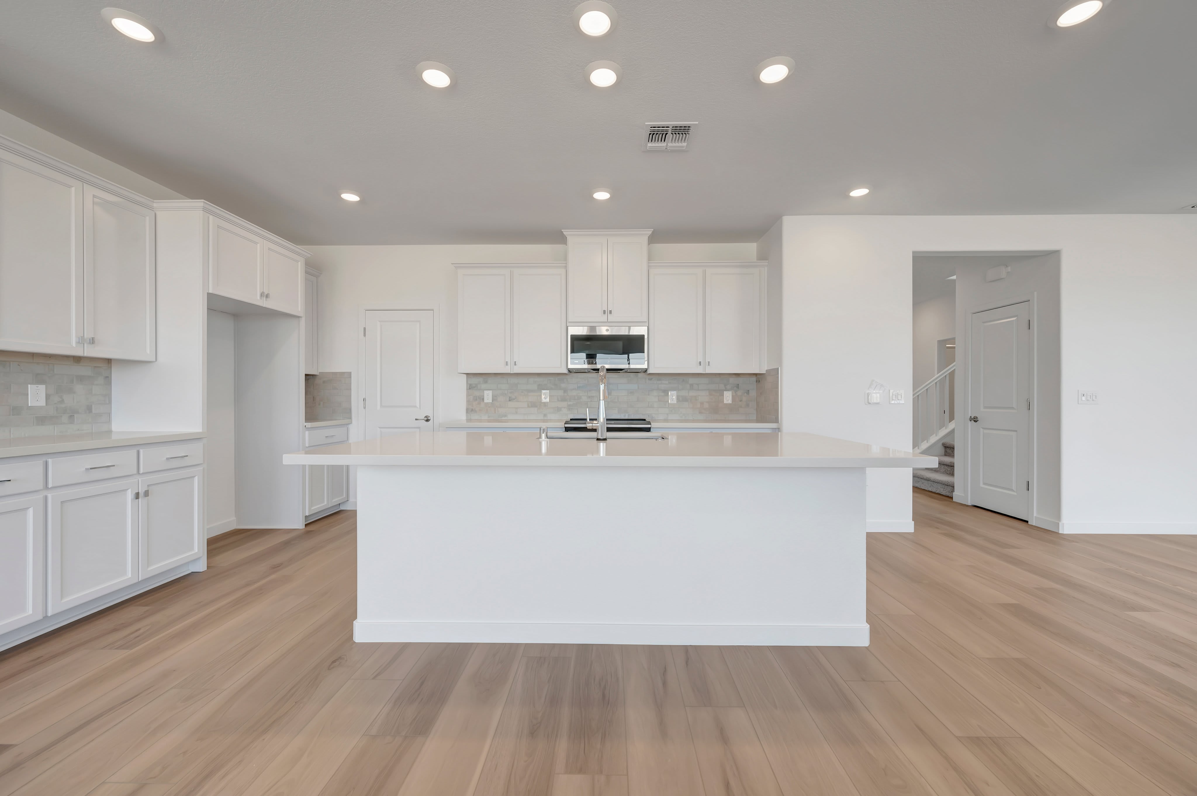 A modern white kitchen with a large island in the foreground and a staircase visible in the background.