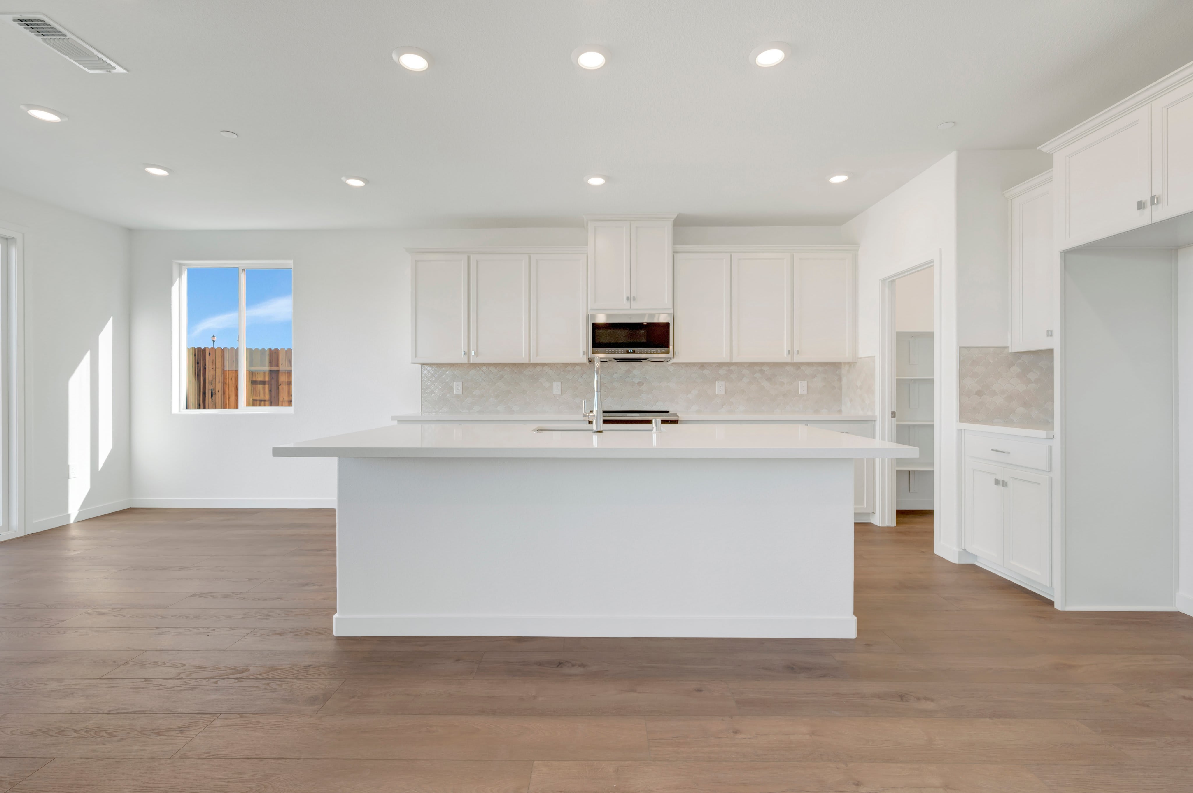 A modern white kitchen with a large island in the foreground is set against a bright living space with a window showing a fence and sky in the background.