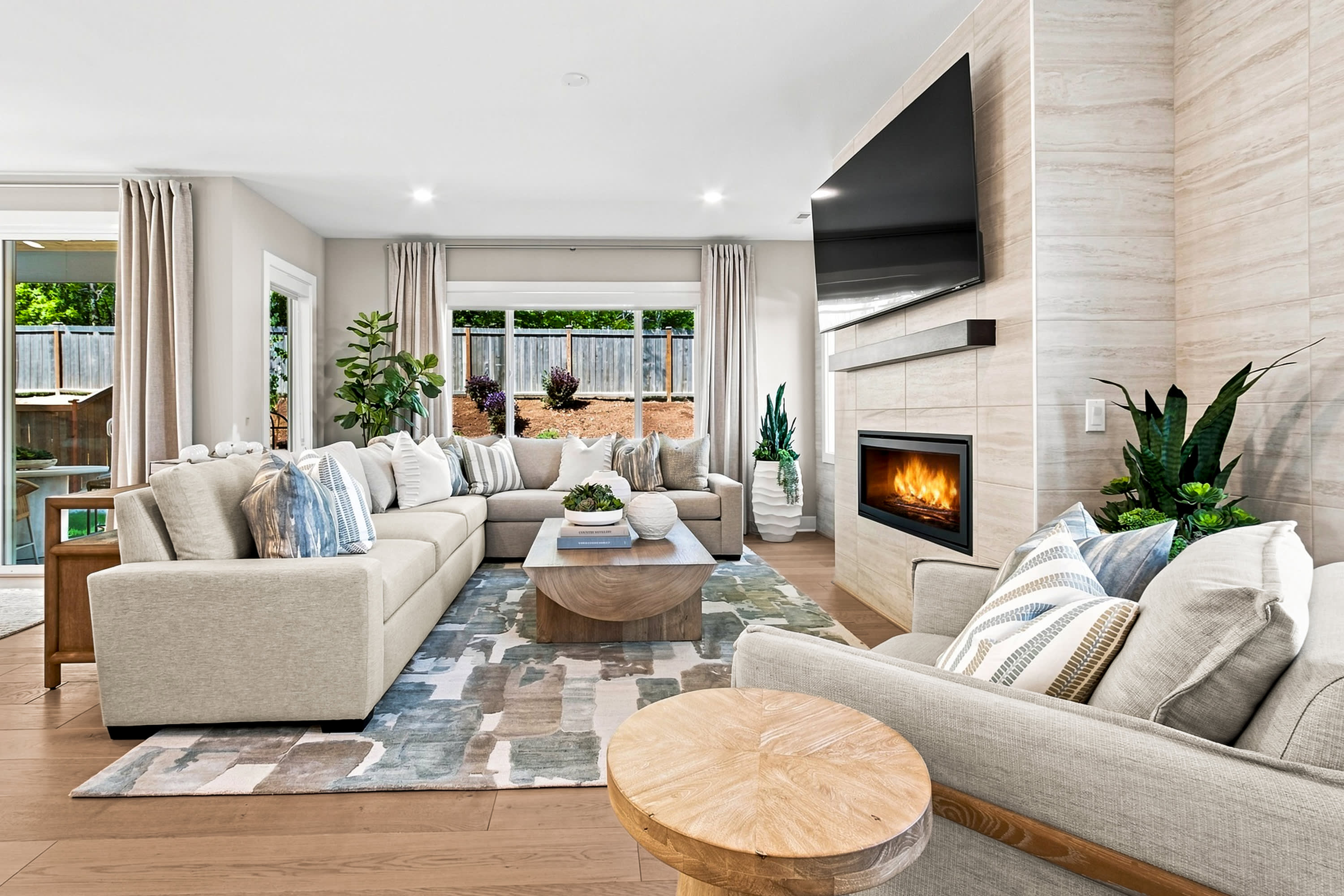 A spacious, modern living room with a large sectional sofa and a fireplace is visible in the foreground, while large windows reveal a backyard with greenery in the background.