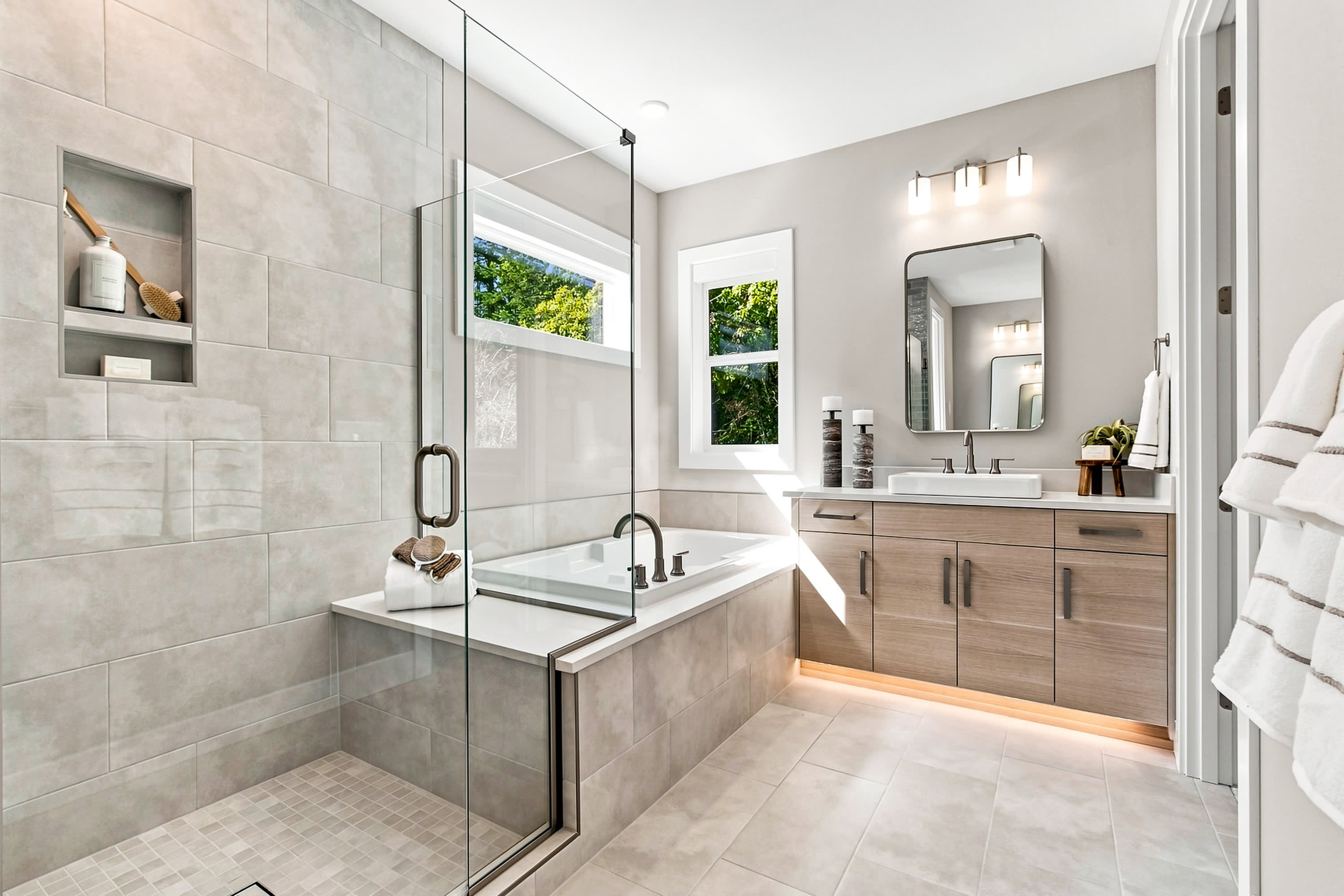 A modern bathroom features a glass-enclosed shower and bathtub in the foreground, with a vanity, sink, and mirror in the background.