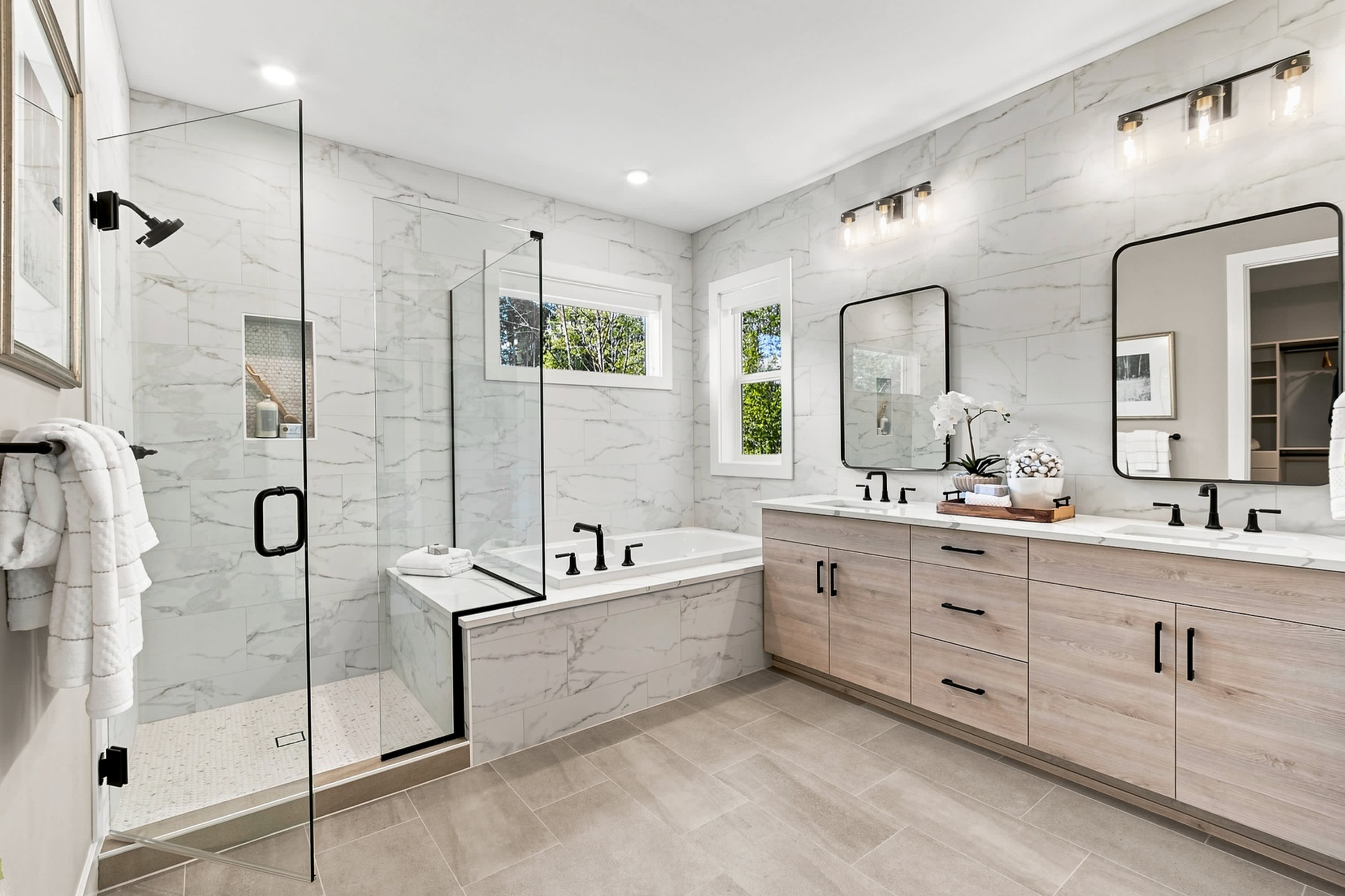 A modern bathroom features a glass-enclosed shower and a freestanding tub in the foreground, with a double vanity and mirrors against a marble-tiled wall in the background.