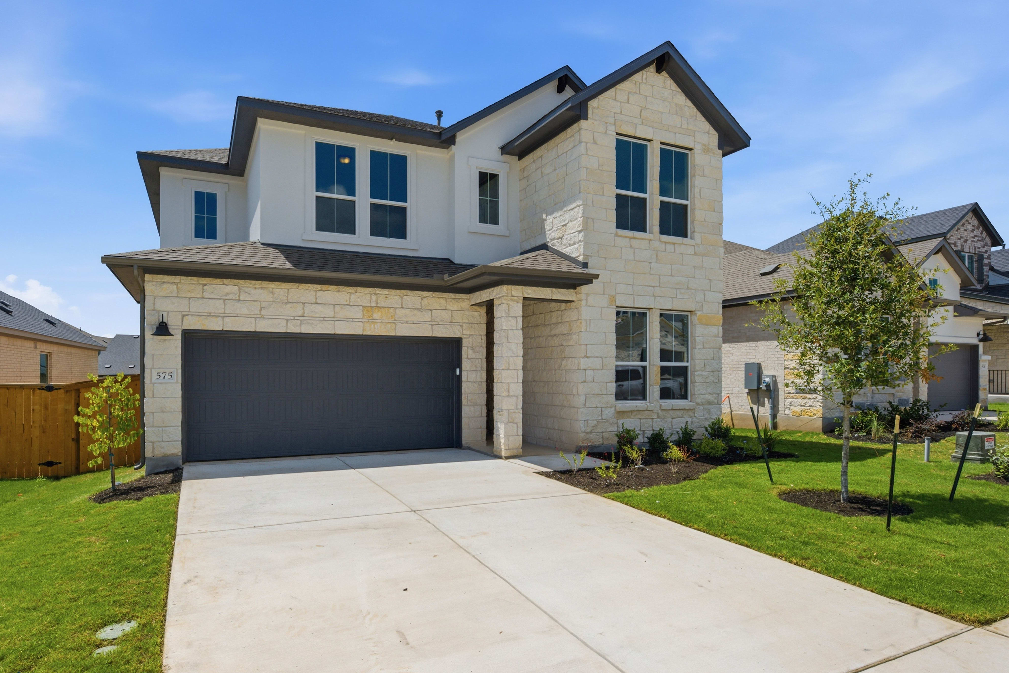 A modern stone-clad house with a dark garage door sits on a manicured lawn in front of a clear blue sky with other houses in the background.