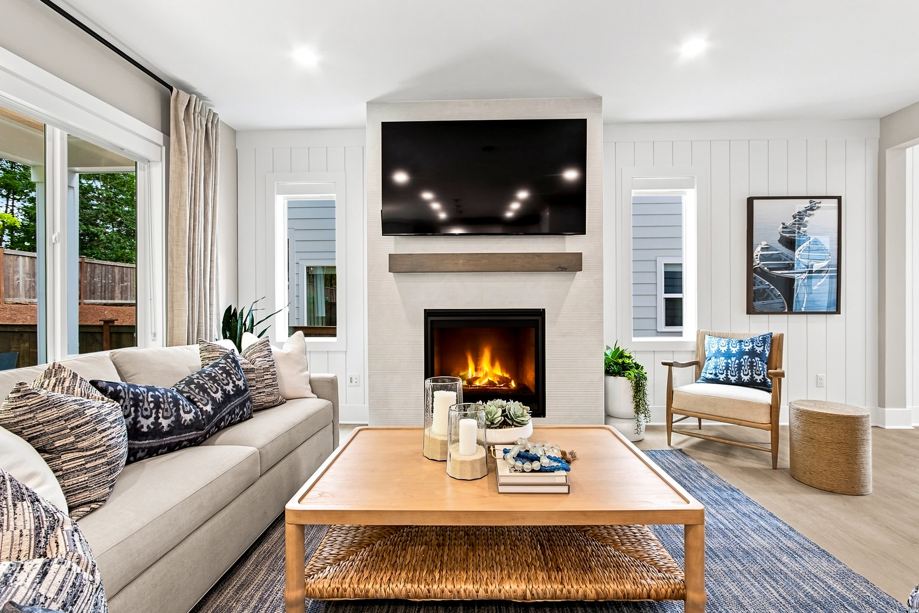 A modern living room features a plush sofa and a wooden coffee table in the foreground, with a fireplace and television as the focal point against a backdrop of windows and a framed artwork.