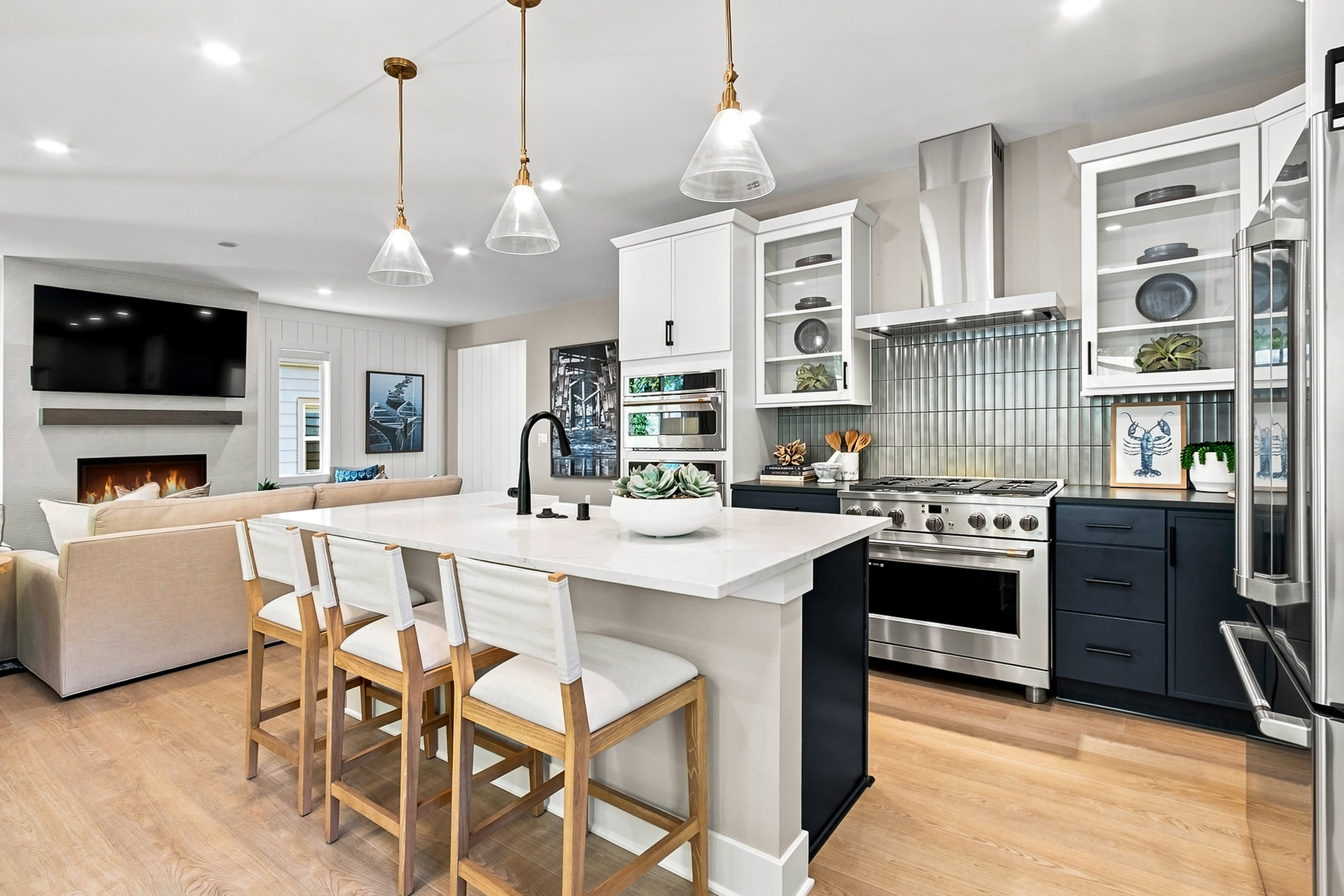 A modern kitchen island with bar stools in the foreground is adjacent to a living area with a fireplace and television in the background.