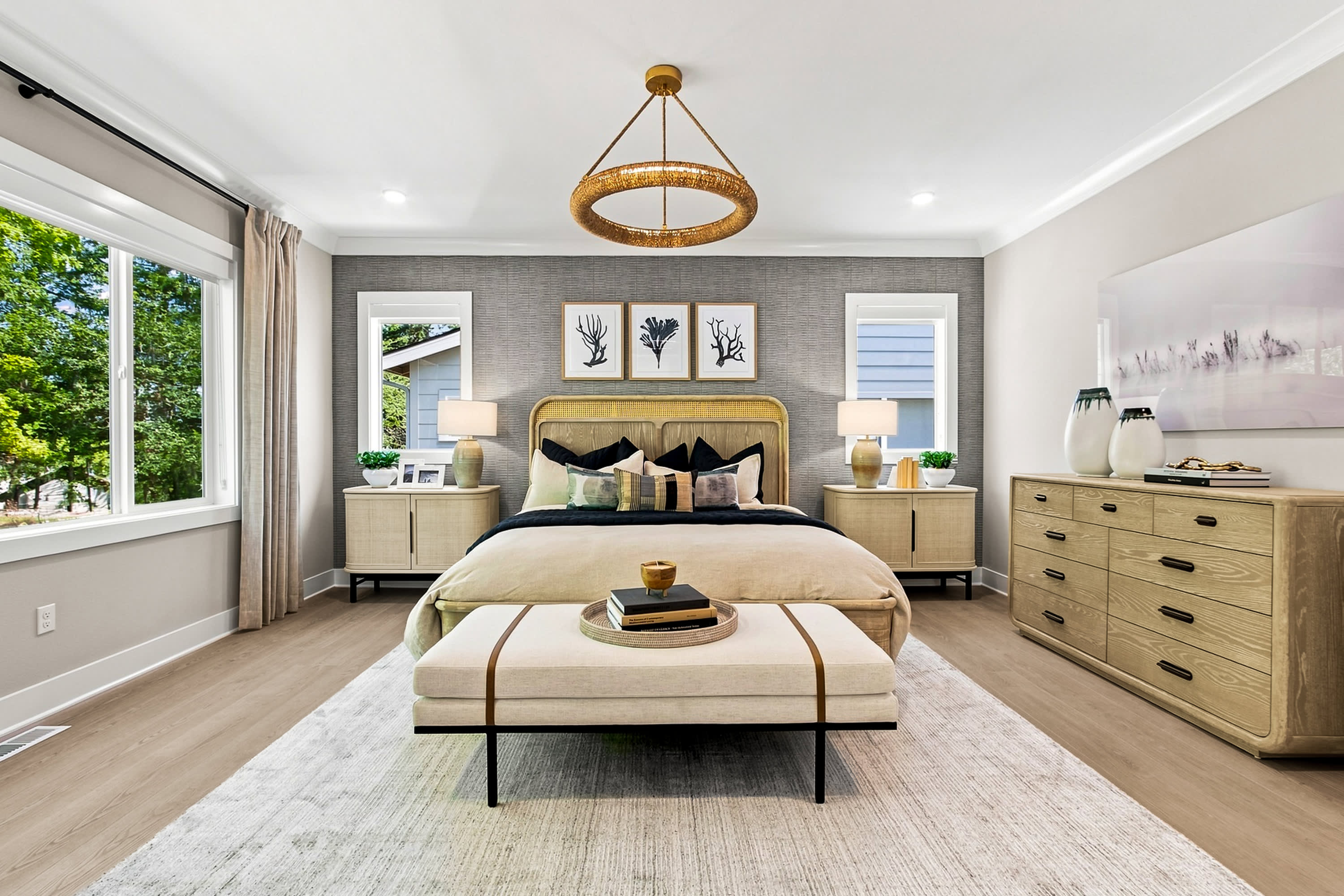 A luxurious bedroom with a king-sized bed and a plush bench in the foreground is set against a backdrop of a textured wall with artwork and large windows overlooking lush greenery.