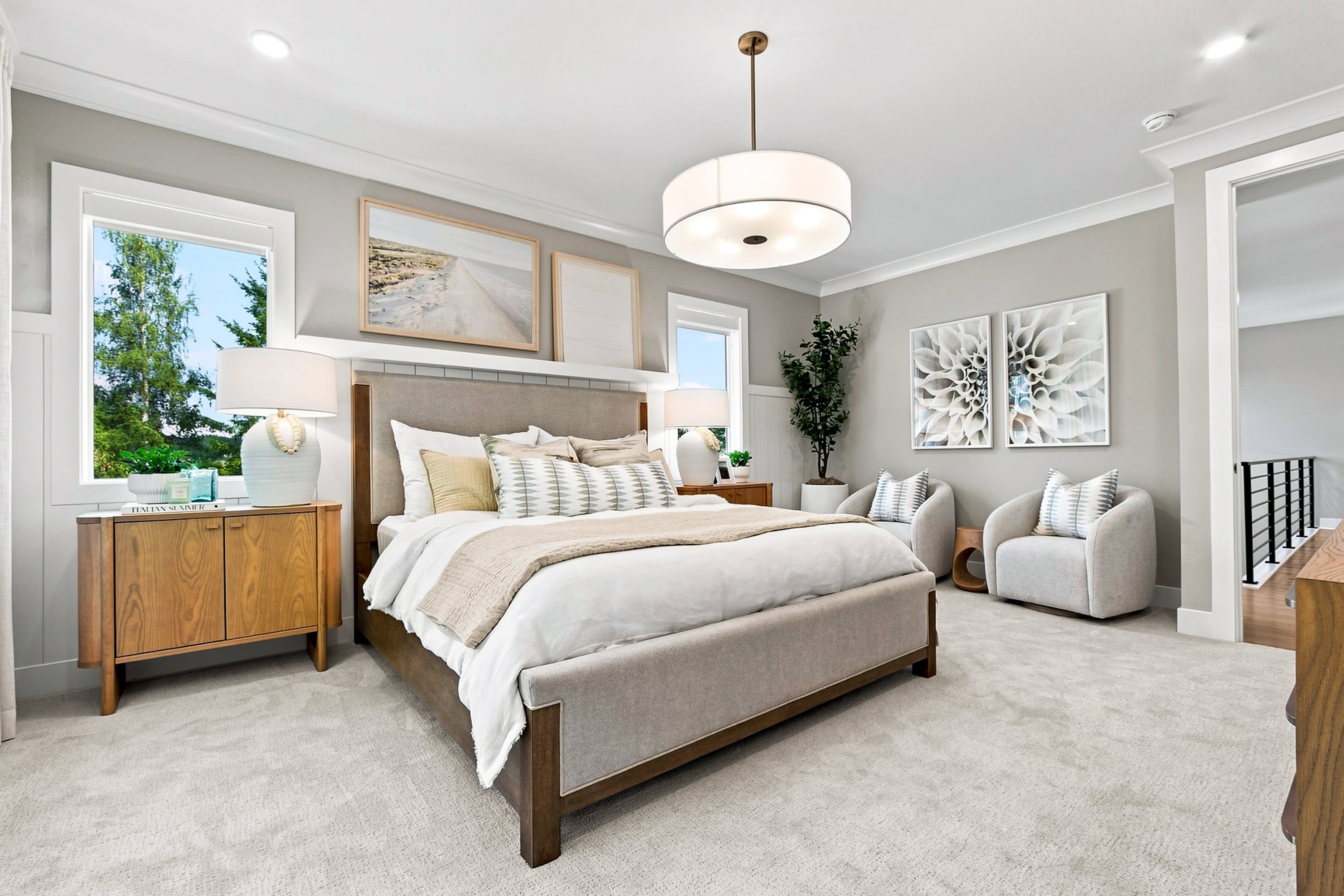 A plush king-sized bed with decorative pillows is centered in a bright bedroom, with two armchairs and artwork on the walls, while a staircase is visible in the background.