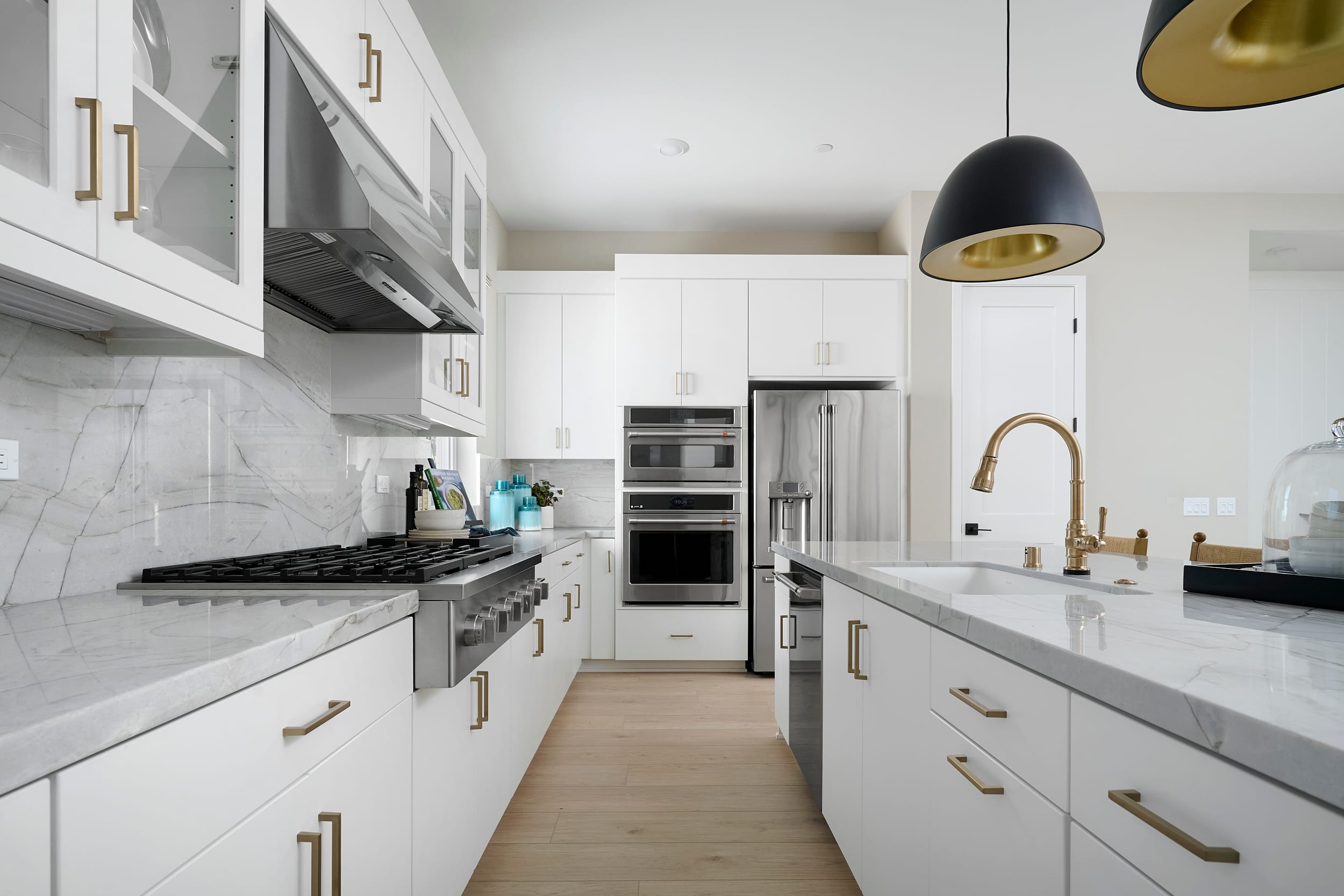 A modern white kitchen with marble countertops and brass fixtures features a sleek gas range in the foreground and stainless steel appliances in the background.