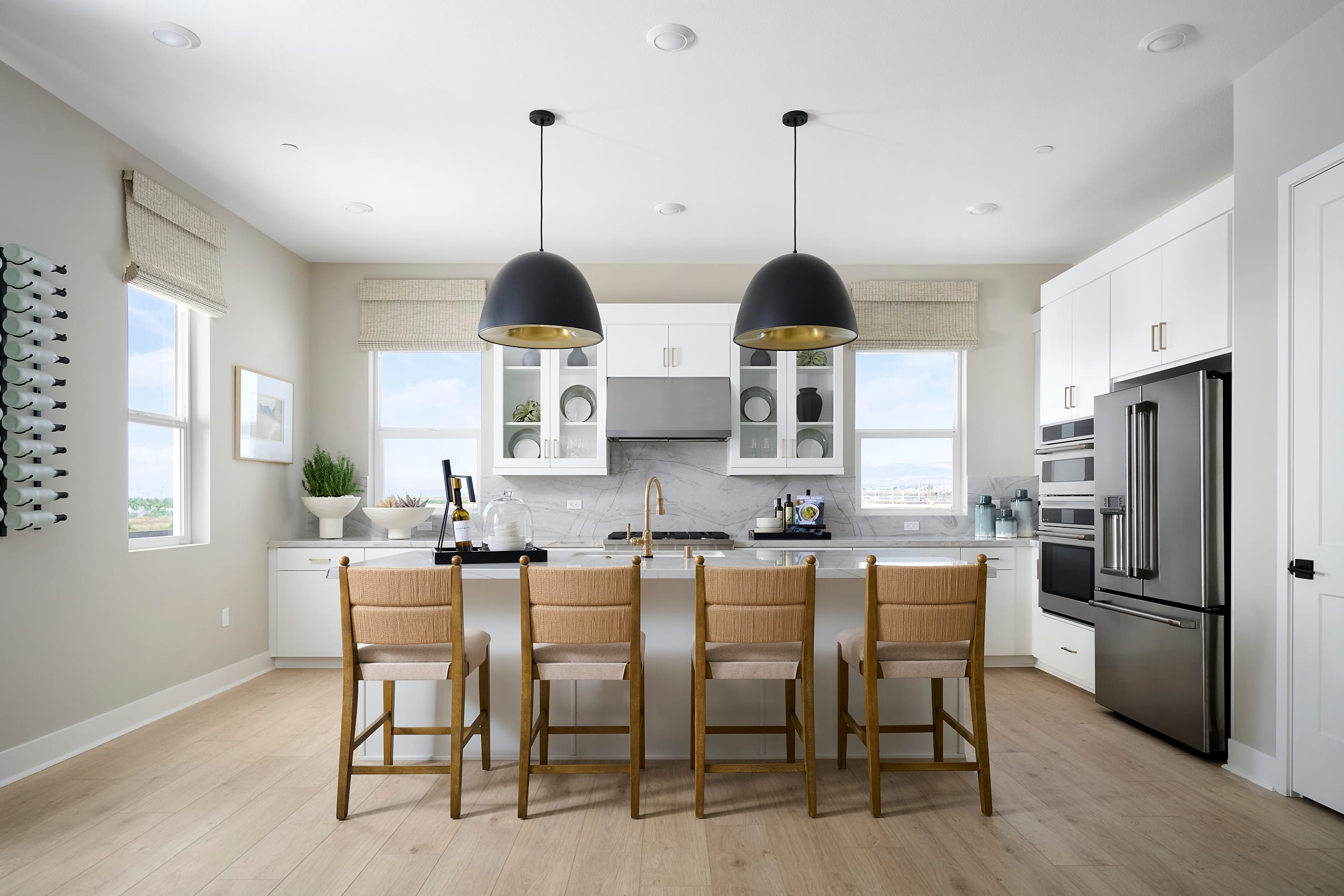 Four woven-seat bar stools are arranged at a white island with a marble countertop, while a stainless steel refrigerator and white cabinets fill the bright, modern kitchen background.