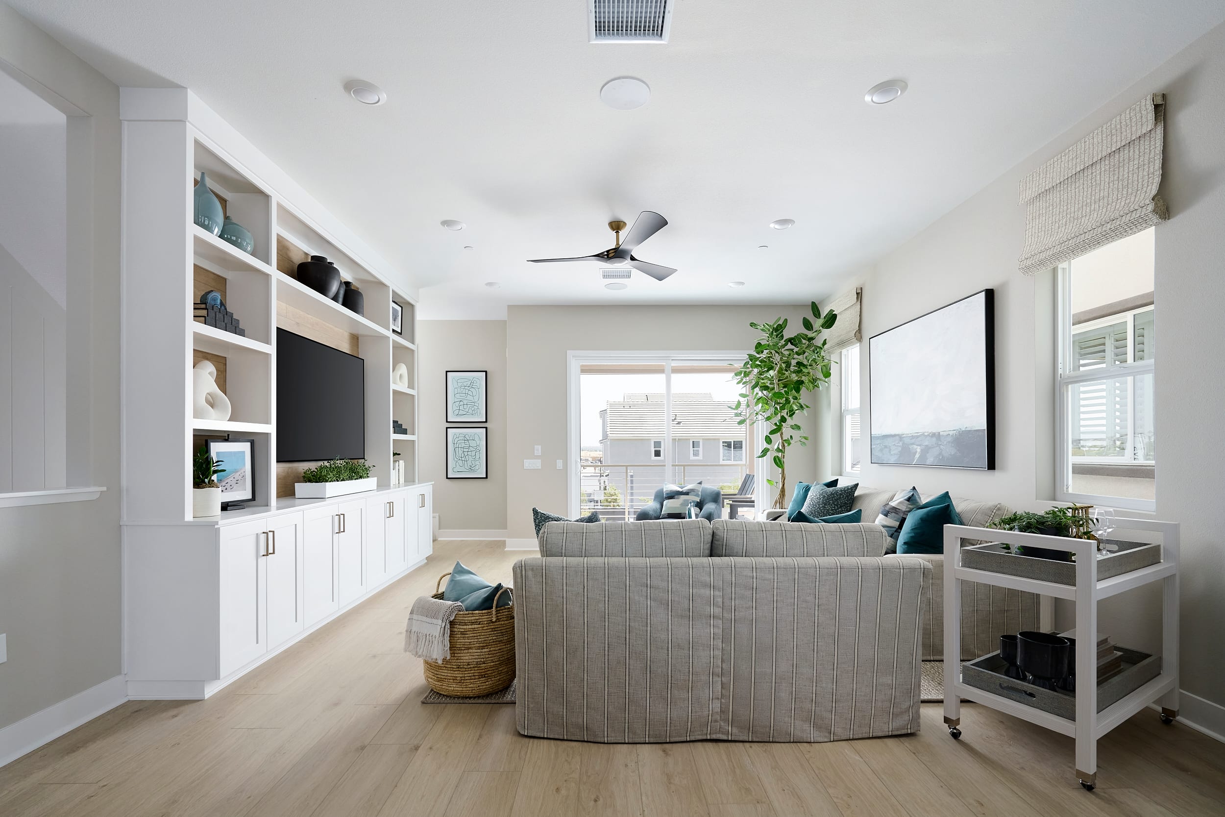 A modern living room with a built-in entertainment center in the foreground and a bright, airy seating area with large windows in the background.