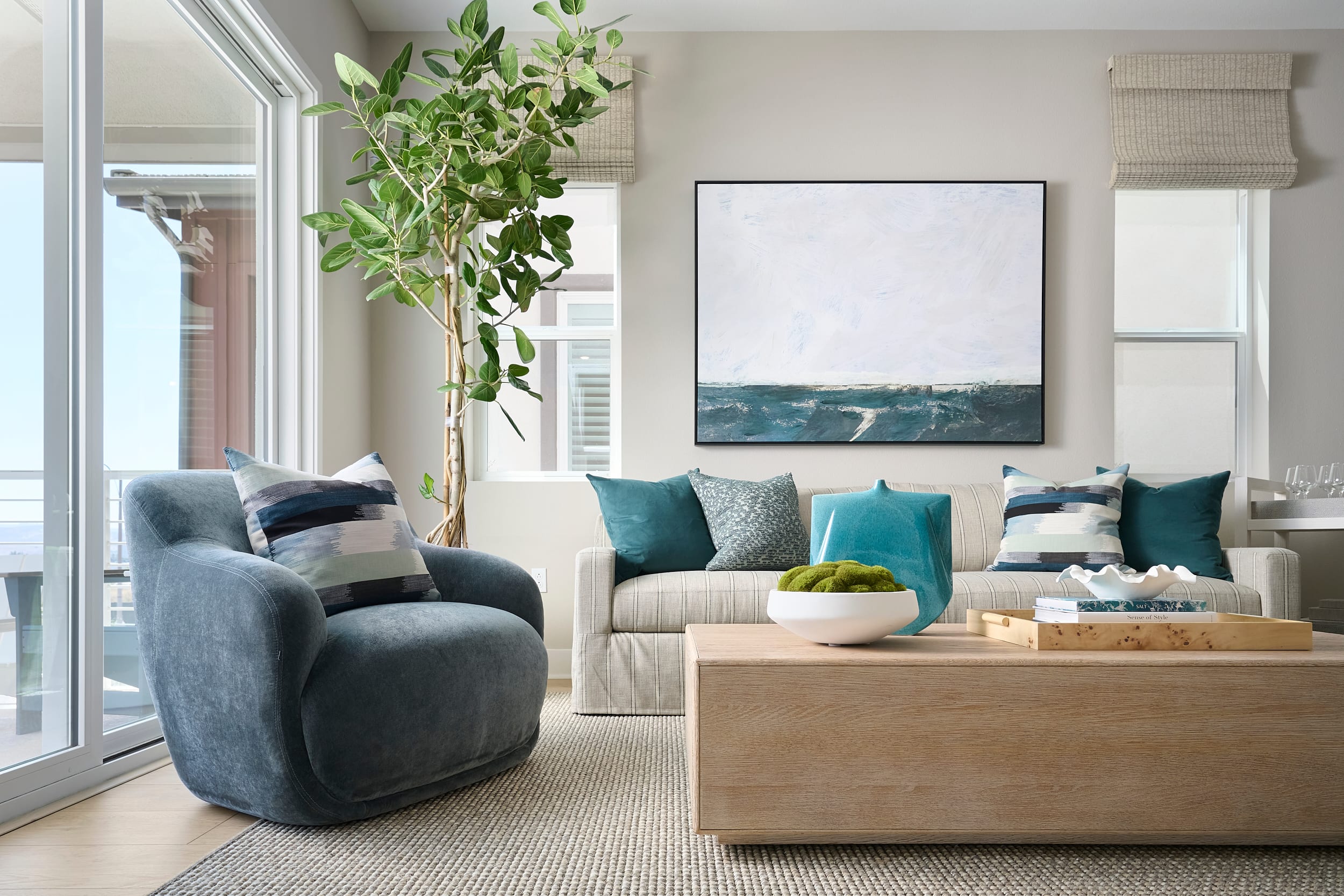 A plush blue armchair and a light wood coffee table with decorative bowls sit on a textured rug in the foreground, while a sofa with teal pillows and a large abstract painting adorns the wall in the background.