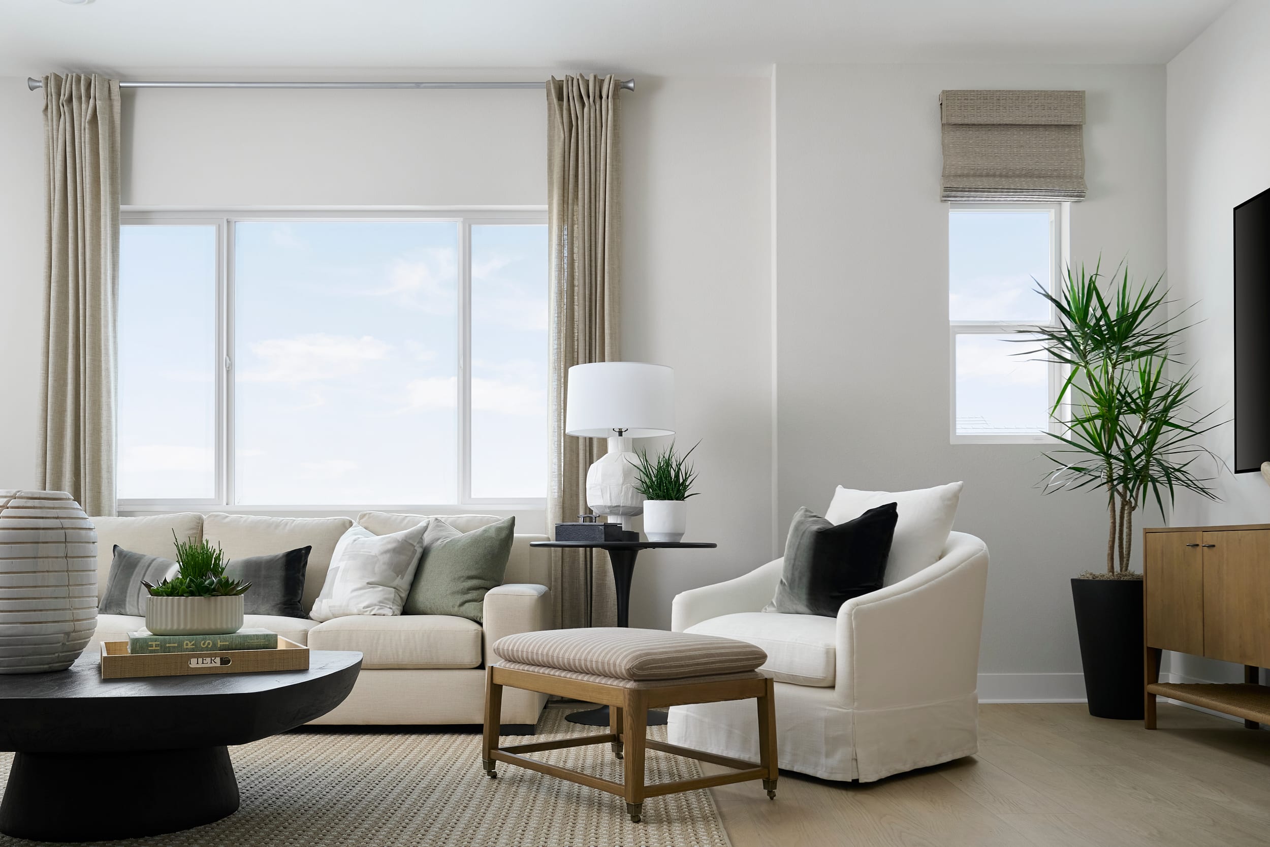 A modern living room with a plush sectional sofa and ottoman in the foreground is bathed in natural light from large windows that reveal a bright sky in the background.