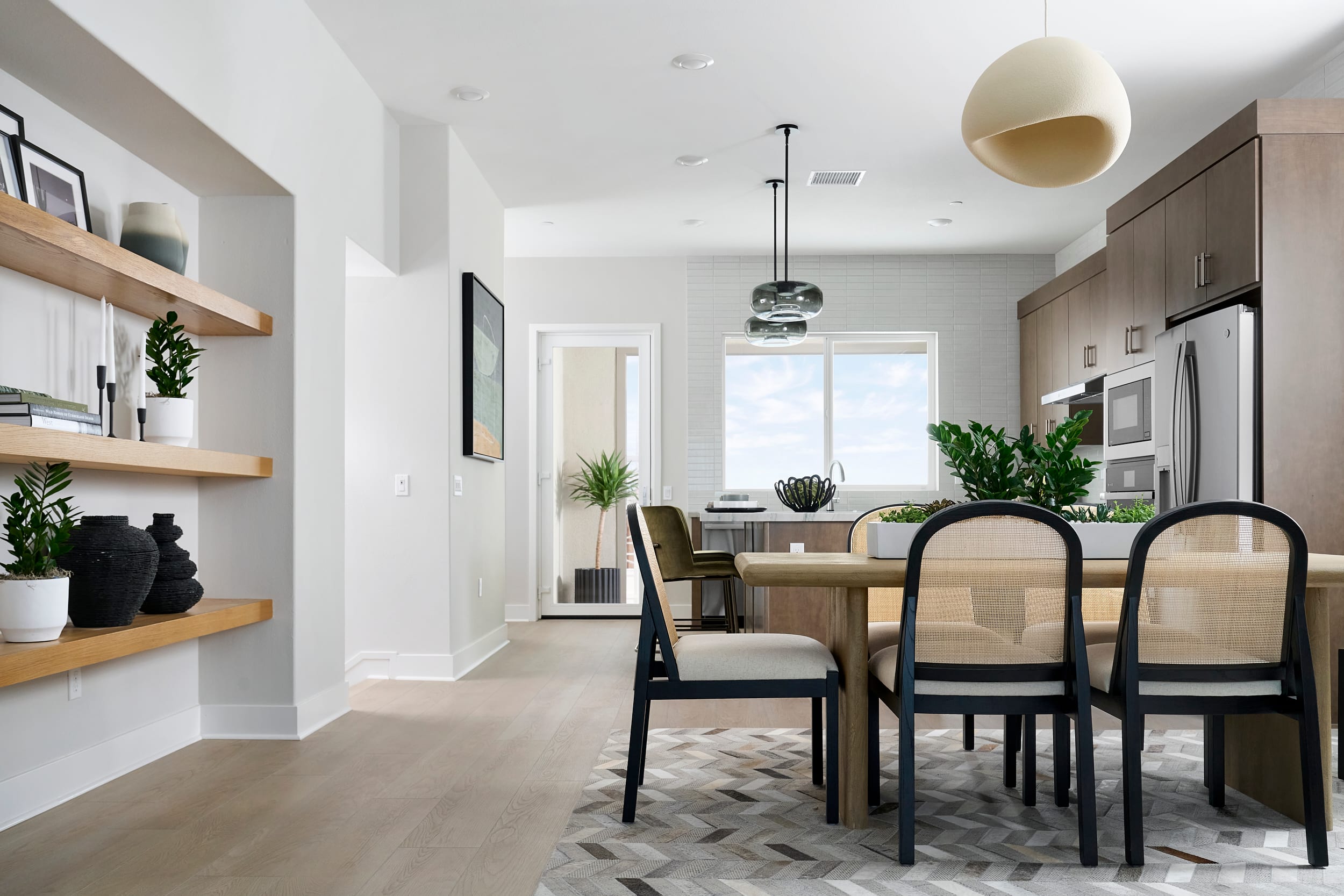 A modern dining area with a light wood table and cane-backed chairs in the foreground is set against a bright kitchen with stainless steel appliances and wooden cabinetry in the background.