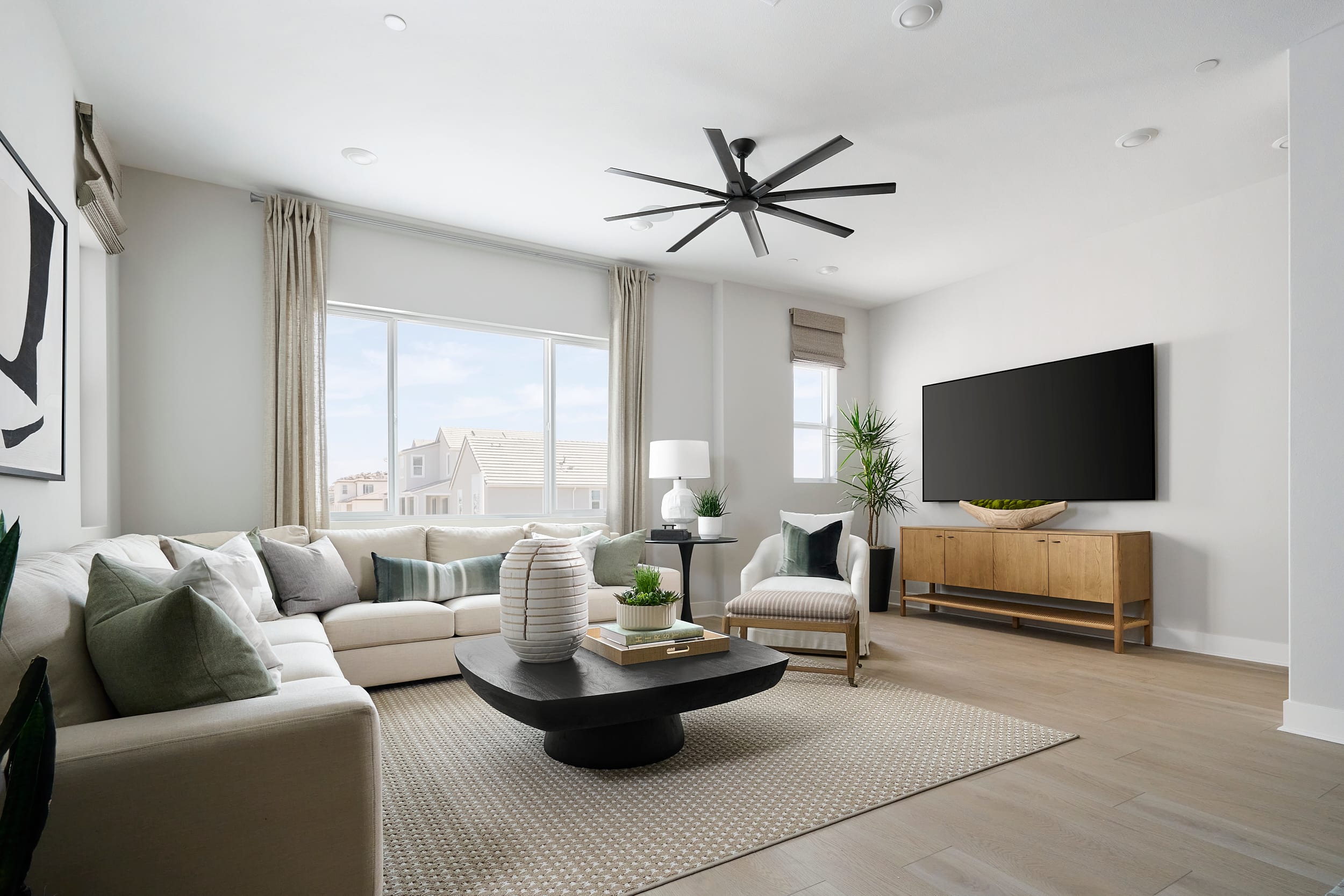 A modern living room with a sectional sofa and coffee table in the foreground, and a large television and window with a view of houses in the background.