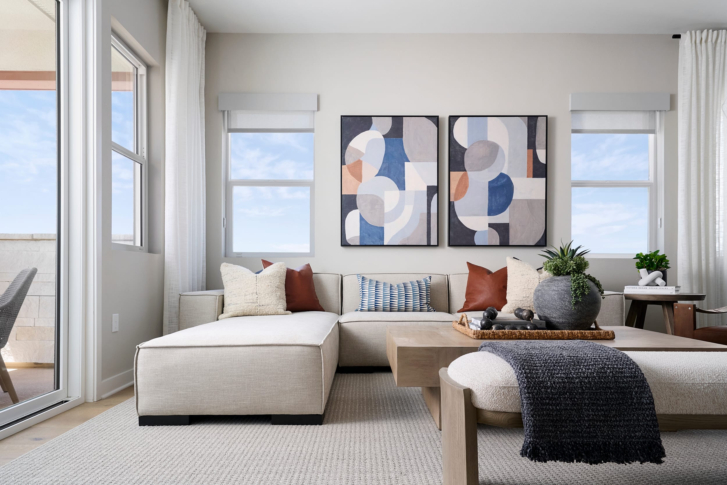 A modern living room with a sectional sofa and coffee table in the foreground is set against a backdrop of large windows revealing a bright sky.