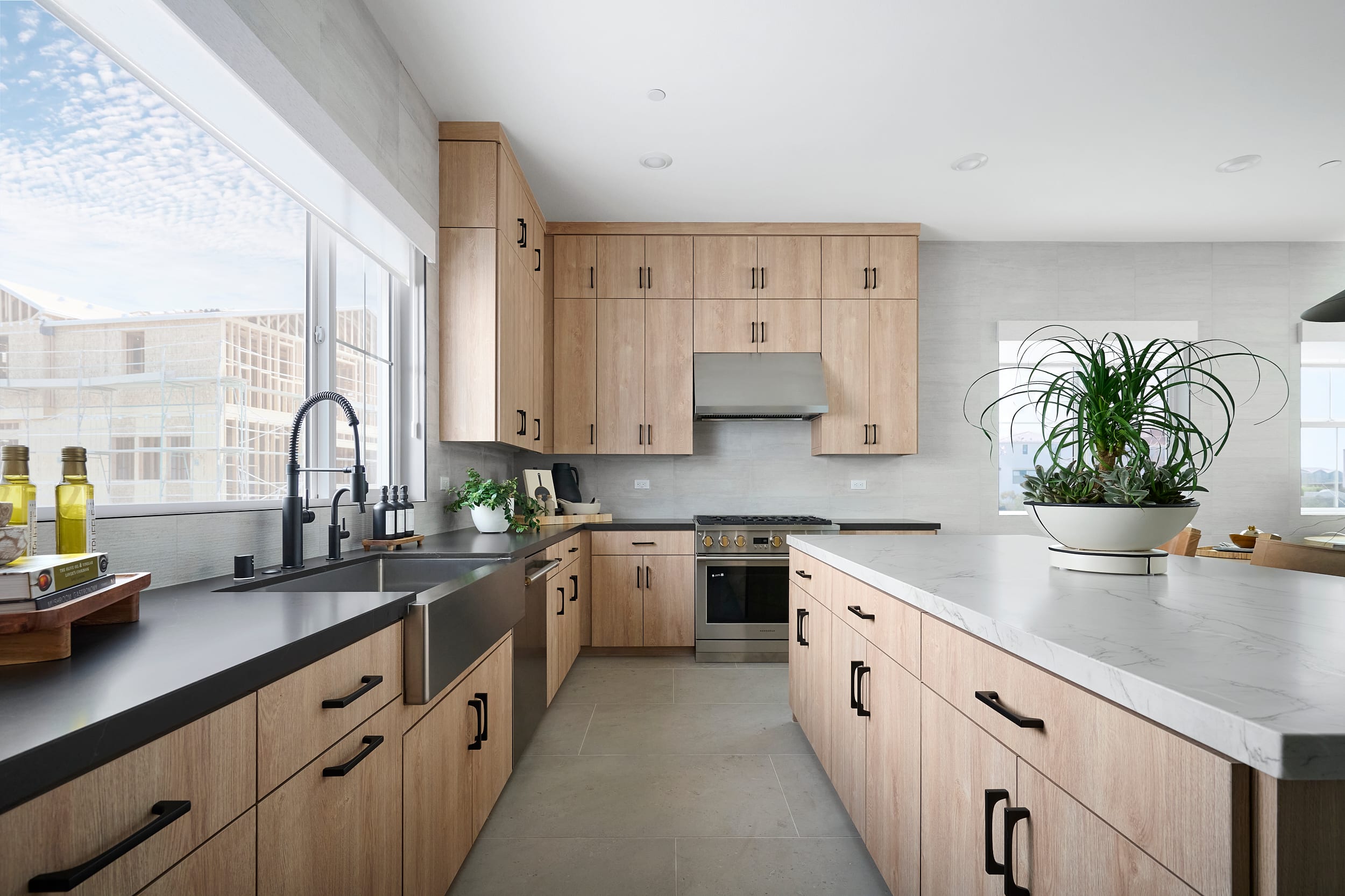 A modern kitchen with light wood cabinets and a large island in the foreground, and a cityscape visible through a large window in the background.