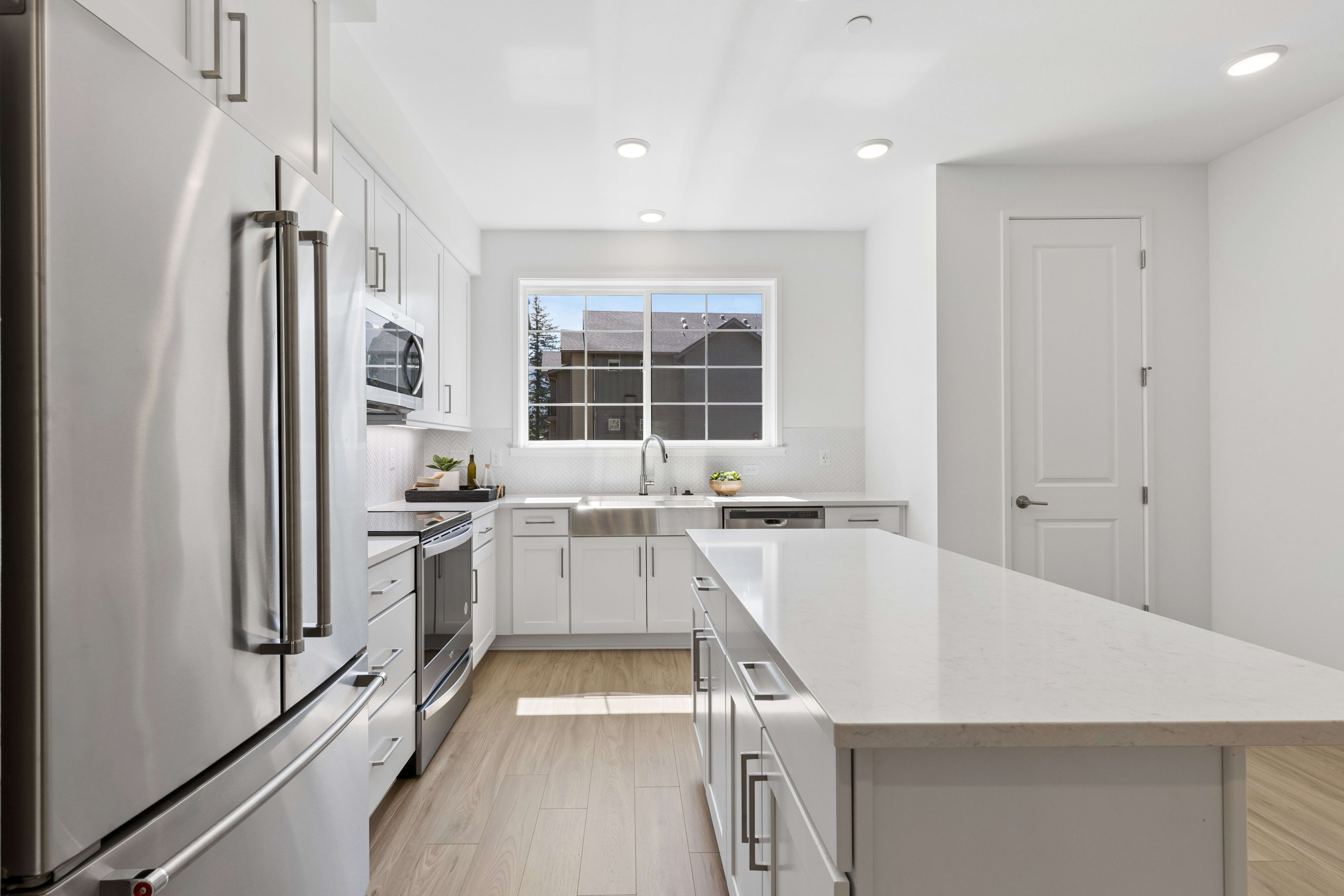 A modern kitchen with a large island in the foreground and a window overlooking houses in the background.