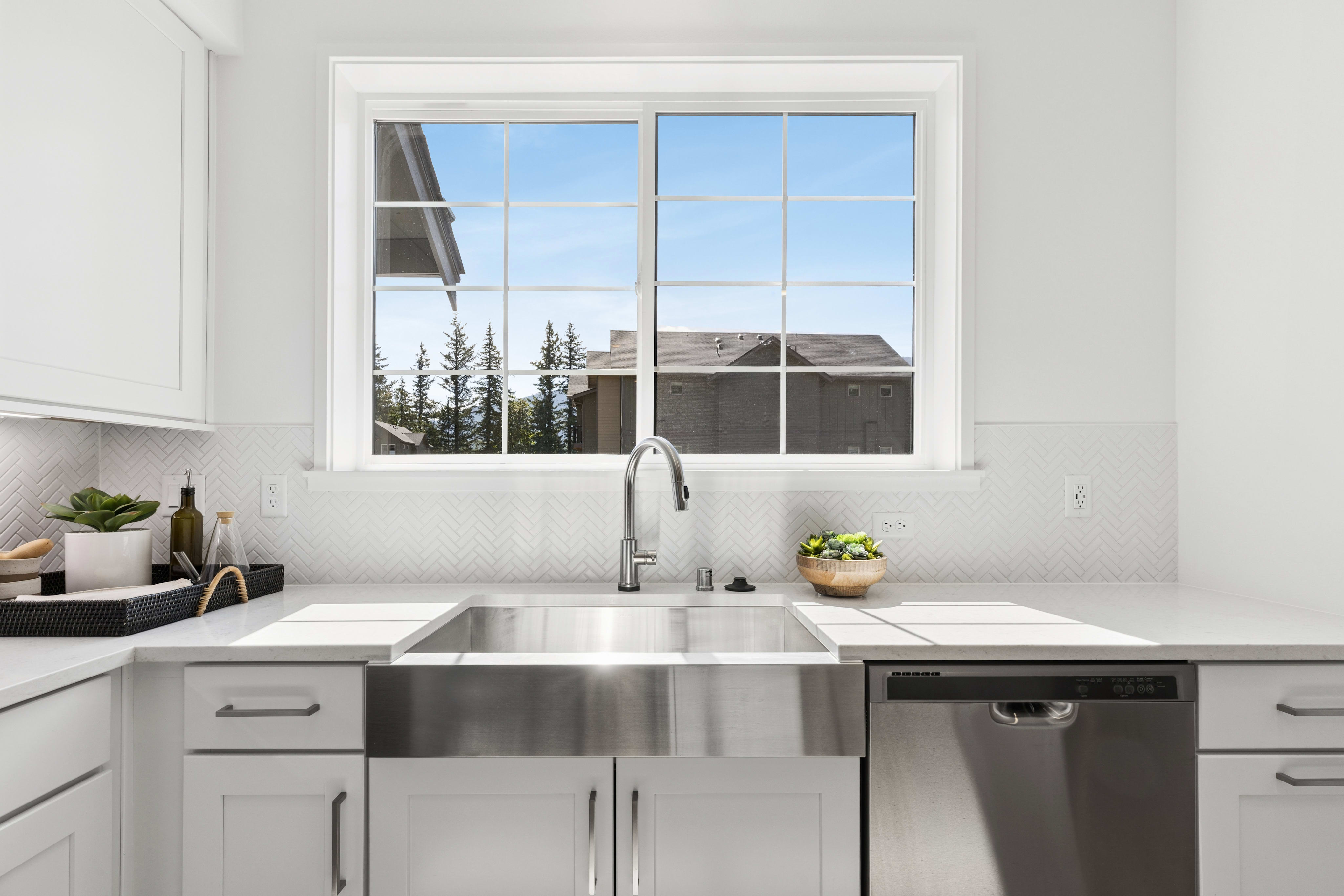 A modern kitchen sink with a stainless steel basin is in the foreground, while a bright window overlooks a suburban neighborhood with trees and houses in the background.