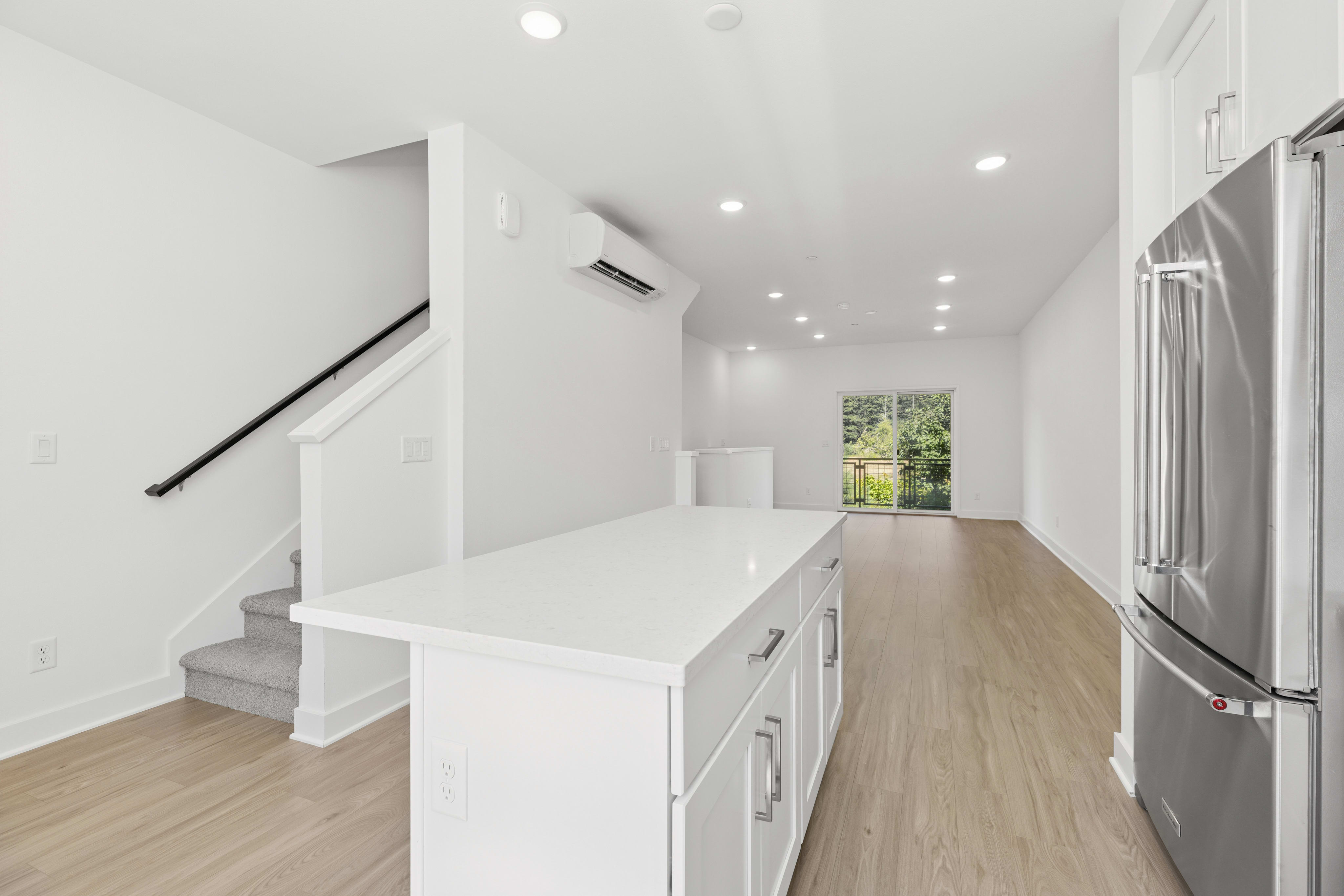 A modern kitchen island with white countertops and cabinets is in the foreground, while a spacious living area with a sliding glass door leading to an outdoor view is in the background.