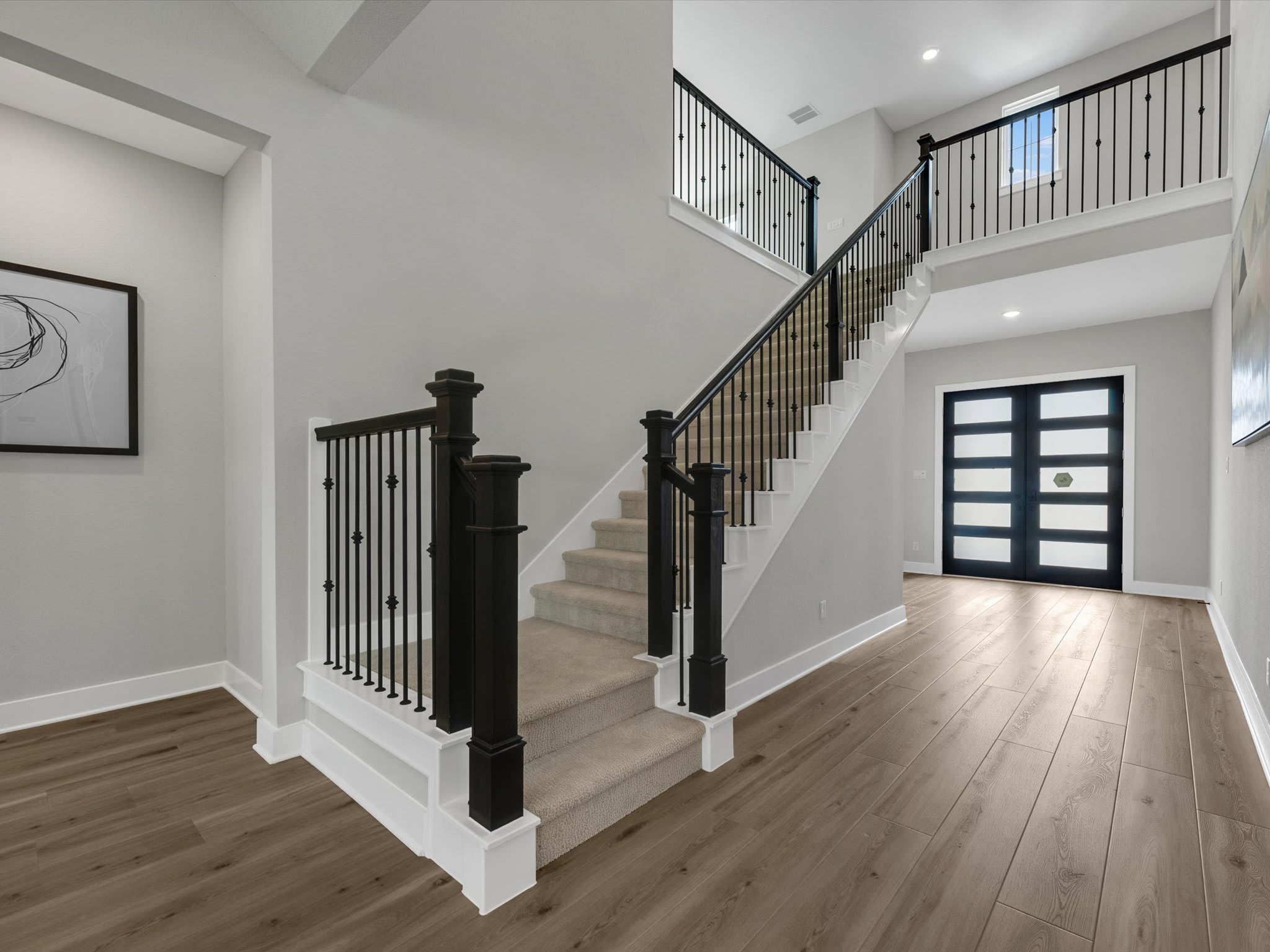 A carpeted staircase with black railings leads up to a landing with a modern black door in the background, while a wood-look floor stretches across the foreground.