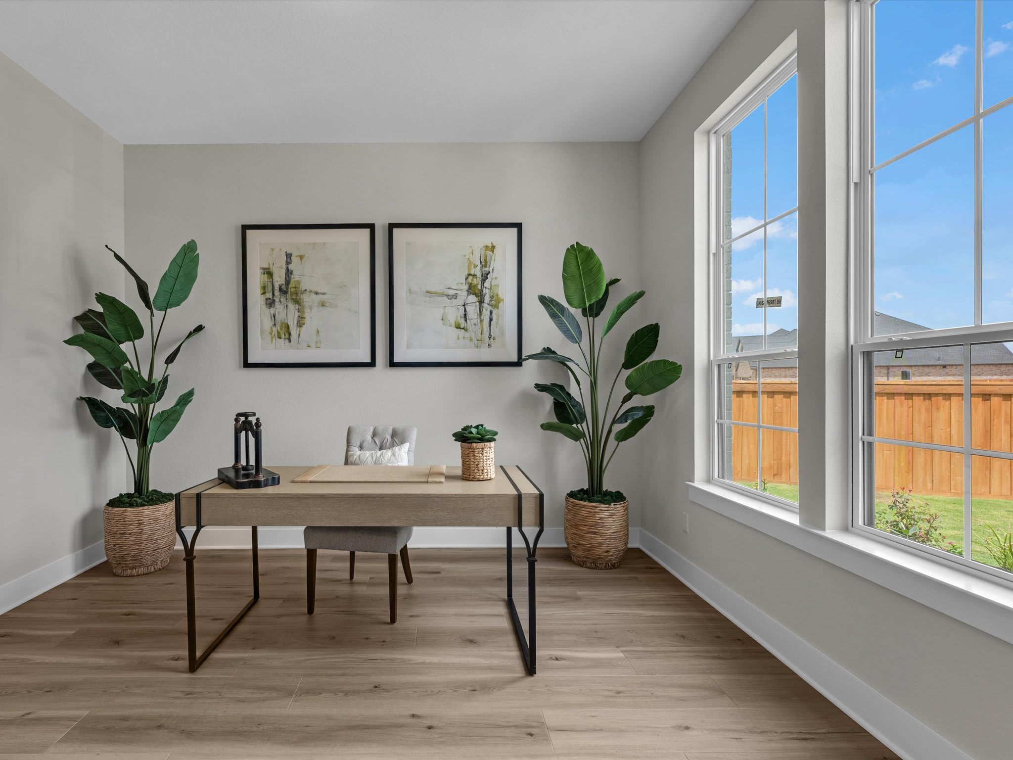 A modern home office with a desk and plants in the foreground is brightly lit by large windows overlooking a backyard with a wooden fence.