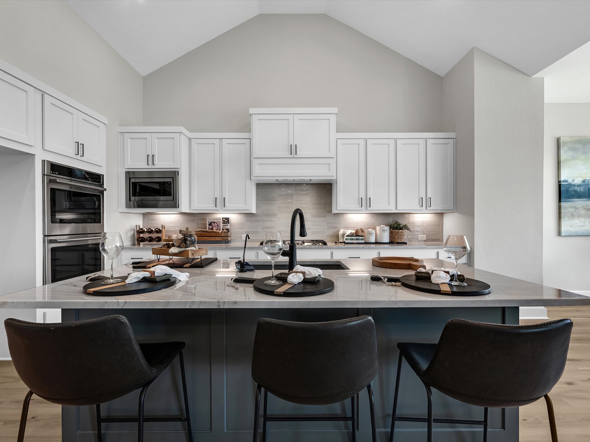 Three dark bar stools sit at a large island in the foreground, while a modern white kitchen with stainless steel appliances and a vaulted ceiling fills the background.