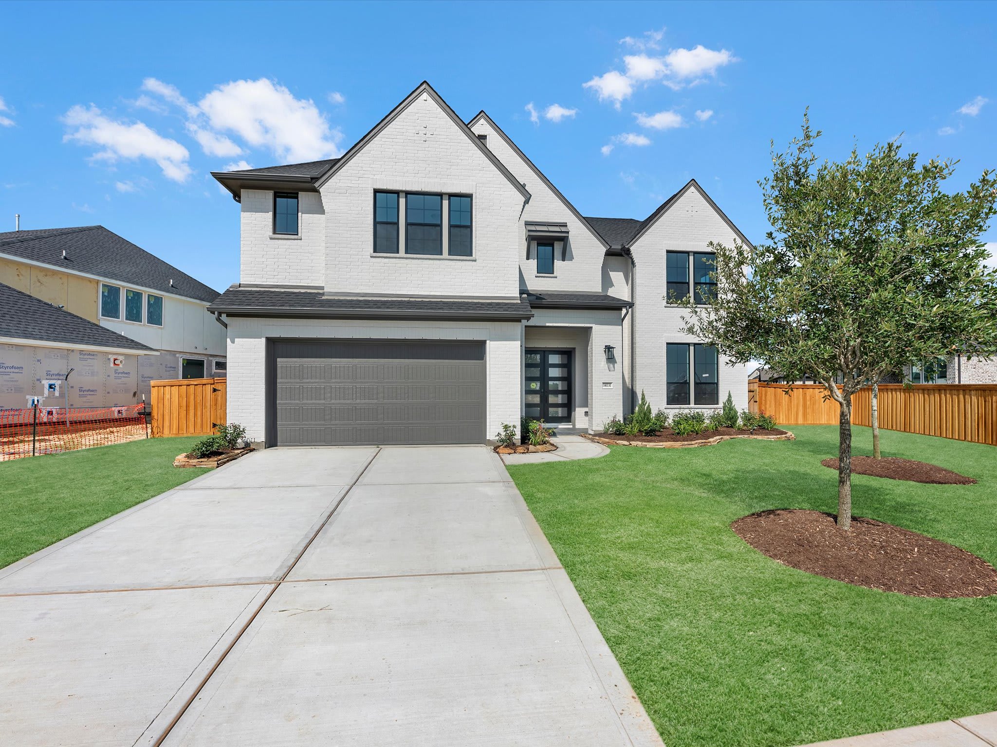 A modern white brick house with a gray garage door sits on a manicured lawn in front of a clear blue sky with scattered clouds and neighboring houses.