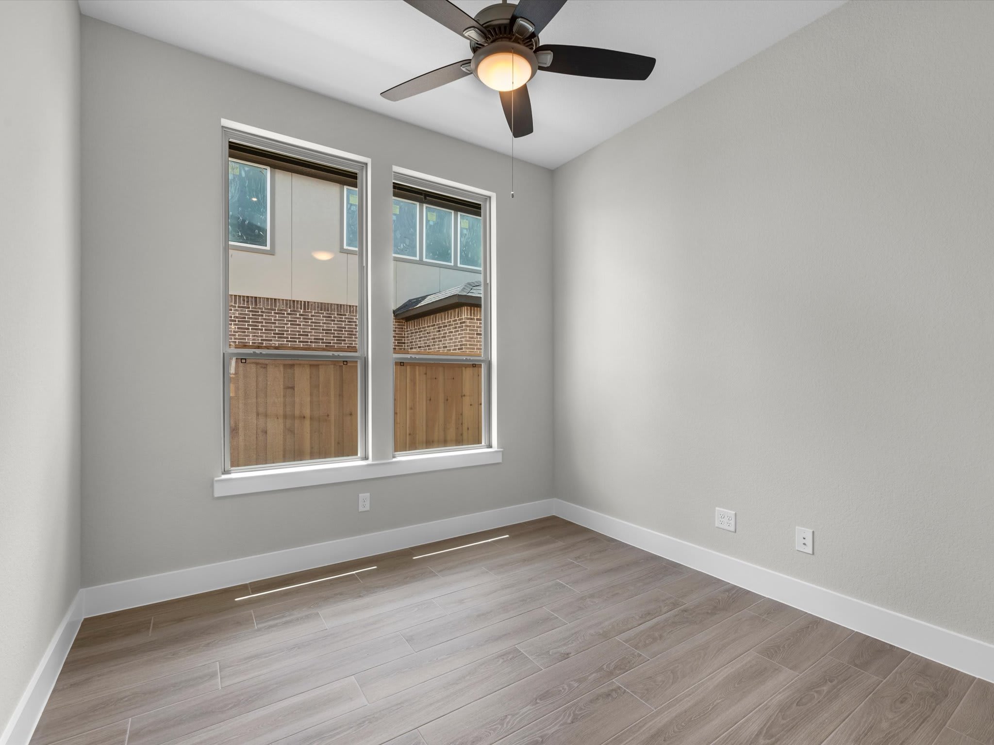 A ceiling fan hangs above a light-colored wooden floor in the foreground, while two large windows in the background offer a view of neighboring houses and a wooden fence.