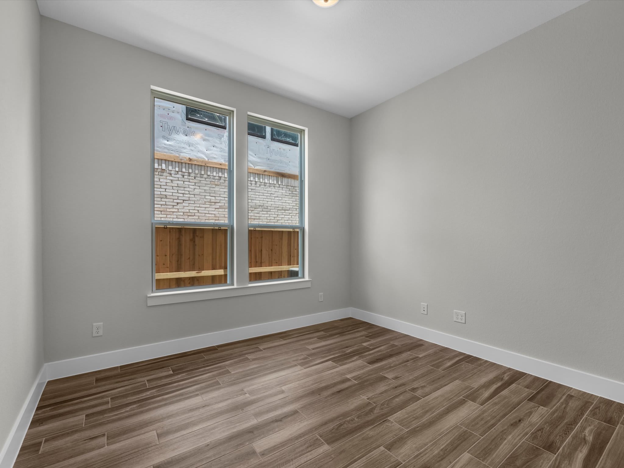 A bright, empty room with wood-look tile flooring in the foreground and two windows revealing a brick wall and wooden fence in the background.