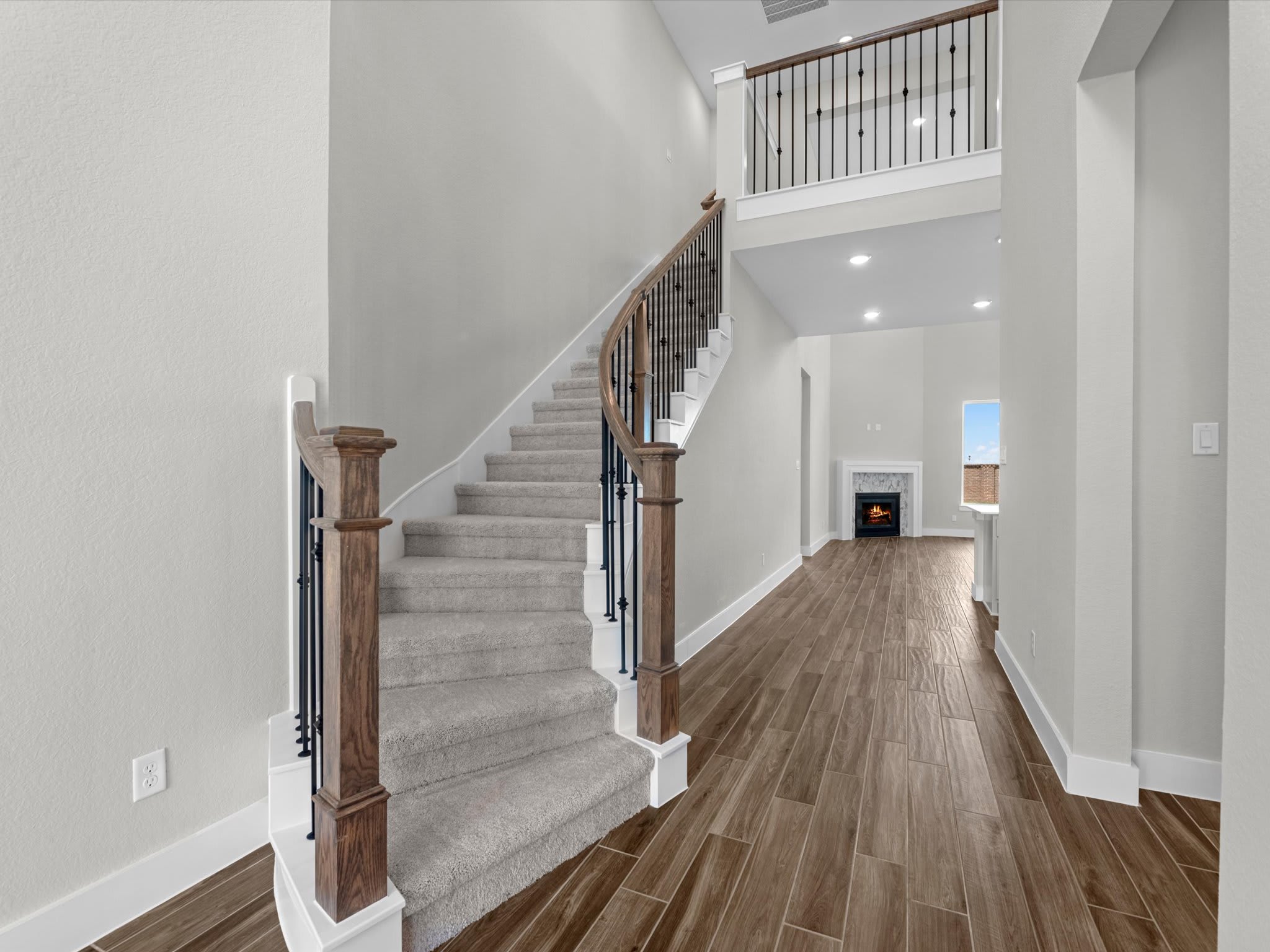 A carpeted staircase with wooden banisters curves upwards in the foreground, leading to a loft area overlooking a living room with a fireplace in the background.
