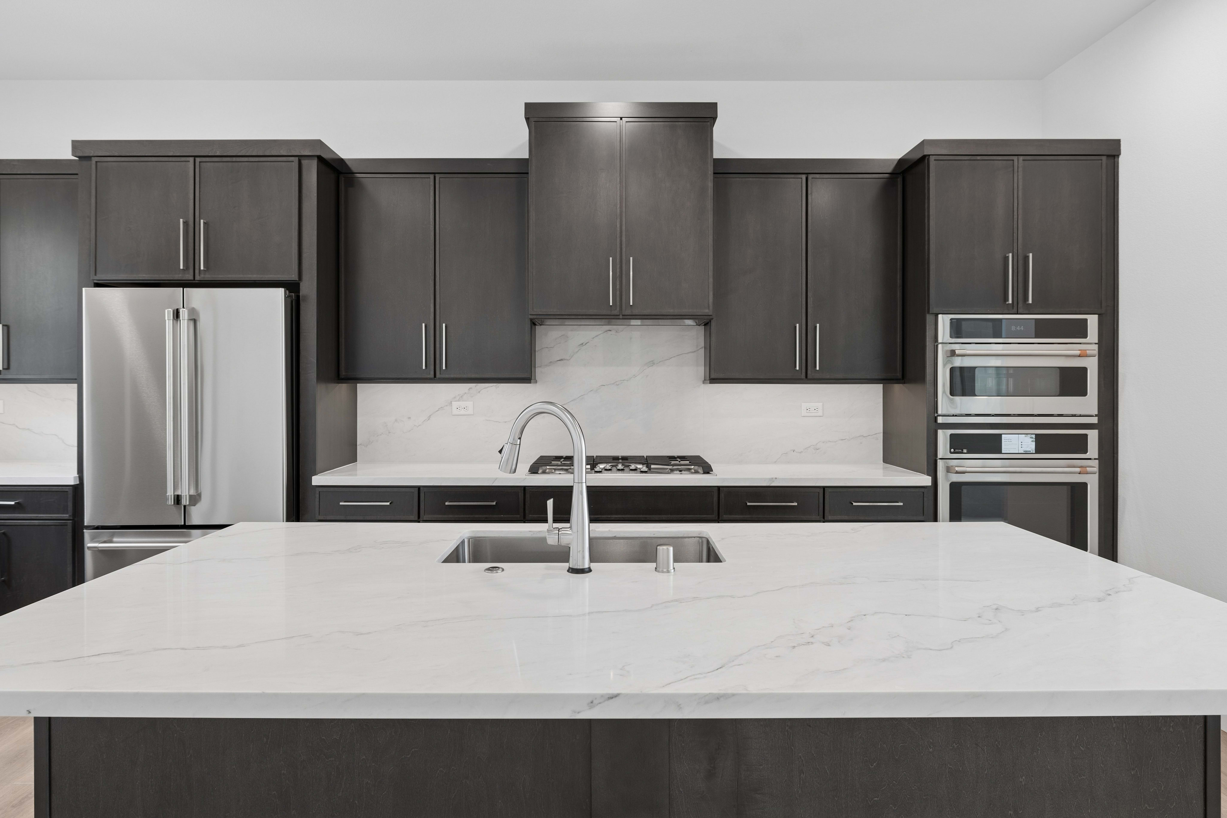 A modern kitchen features a white marble island in the foreground and dark gray cabinetry with stainless steel appliances in the background.