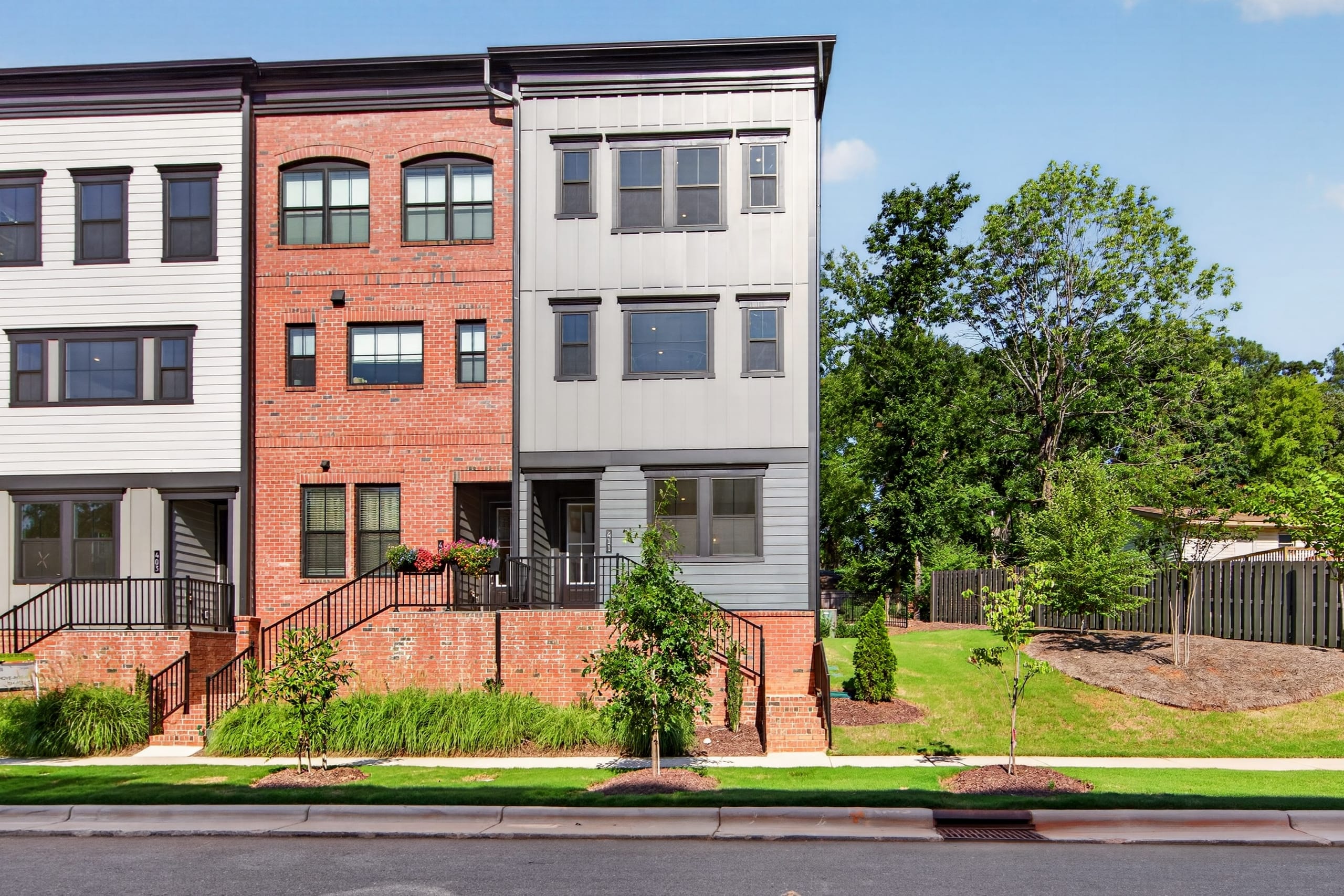 A row of modern townhouses with brick and metal facades stands in the foreground, backed by a lush green lawn and mature trees under a clear blue sky.
