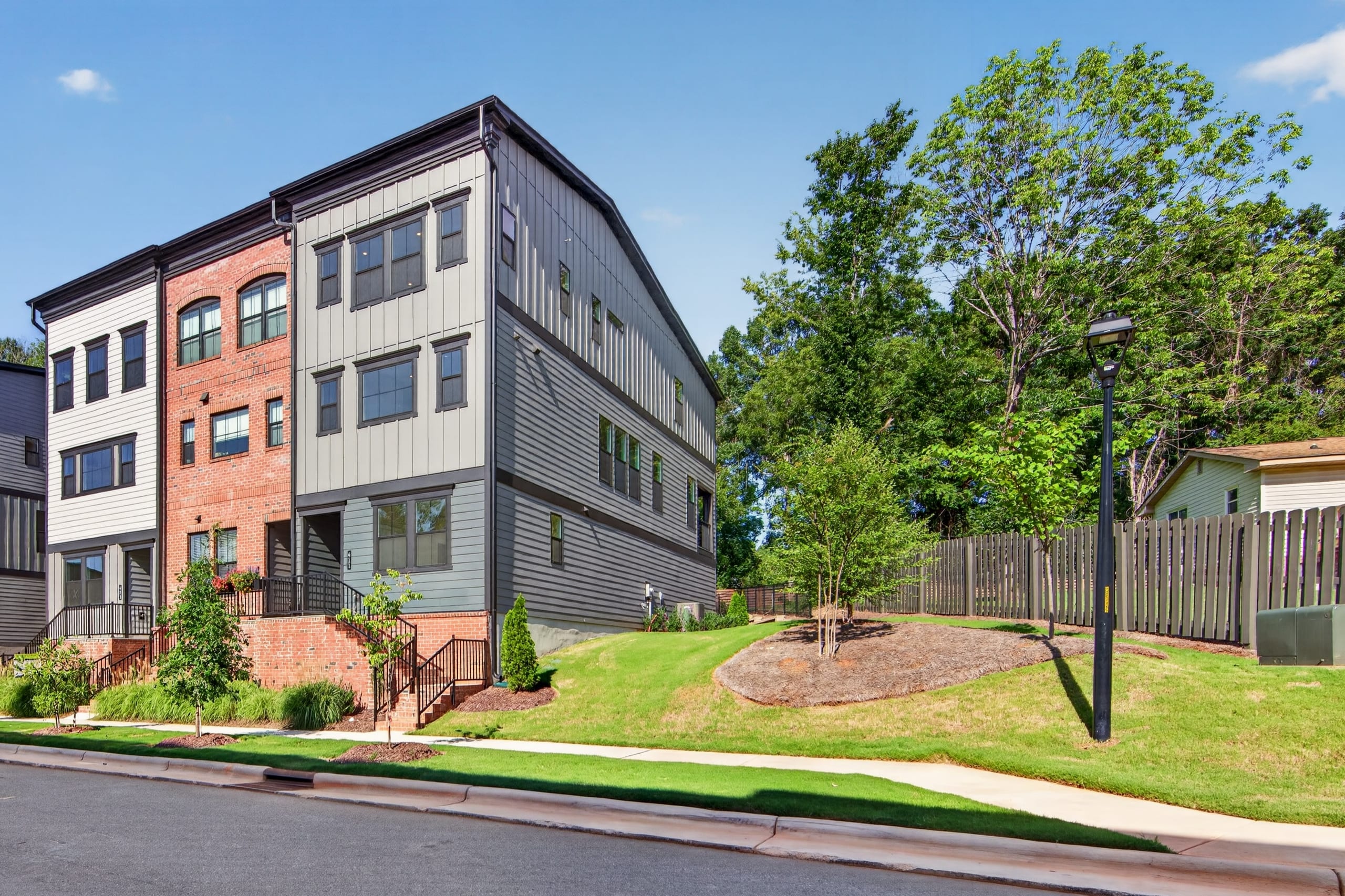 Modern townhouses with brick and gray siding stand on a manicured lawn with a wooden fence and trees in the background.