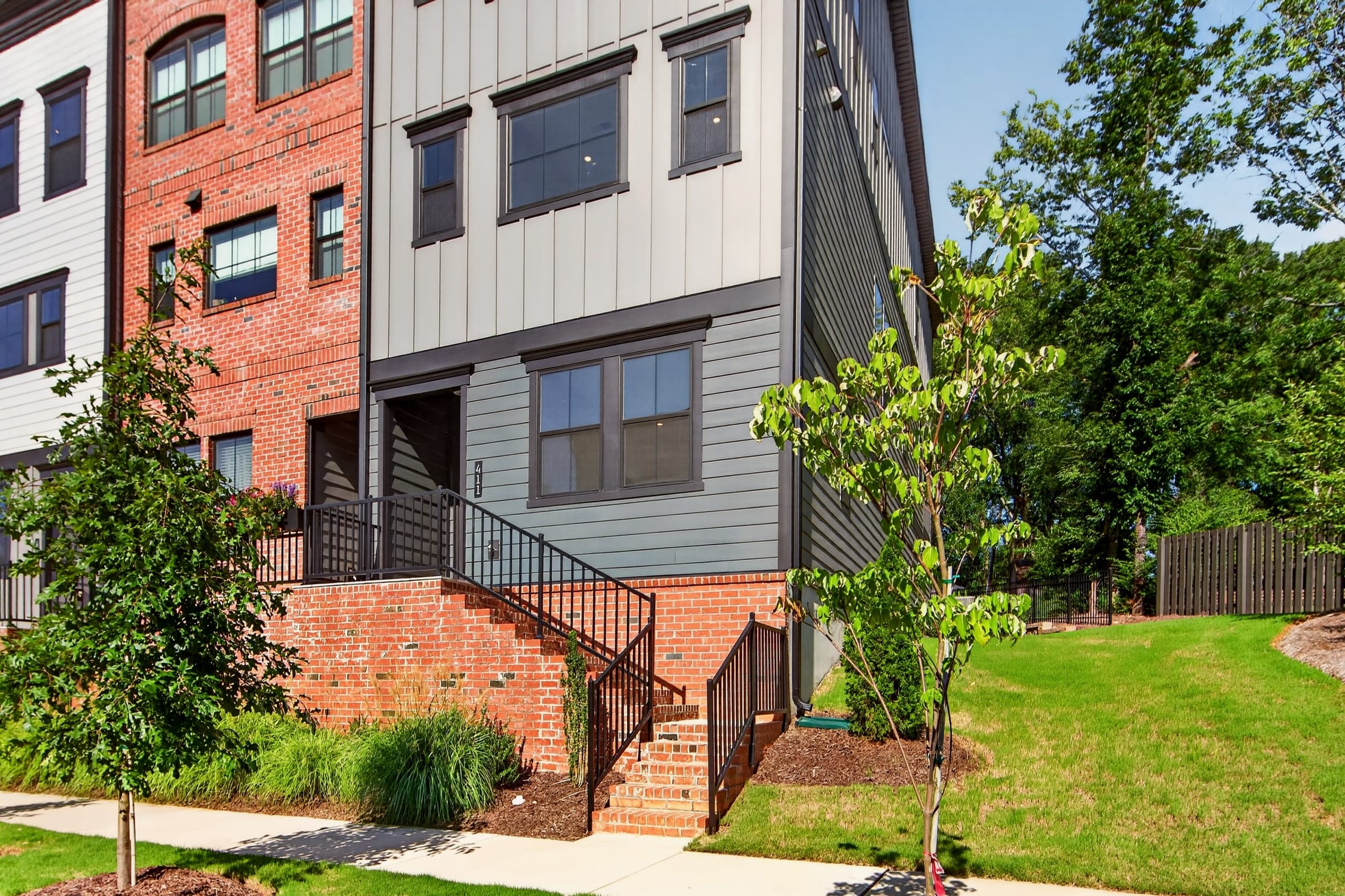 A modern townhouse with a brick and gray siding exterior stands on a grassy lawn with trees in the foreground, while a wooden fence and lush greenery form the background.