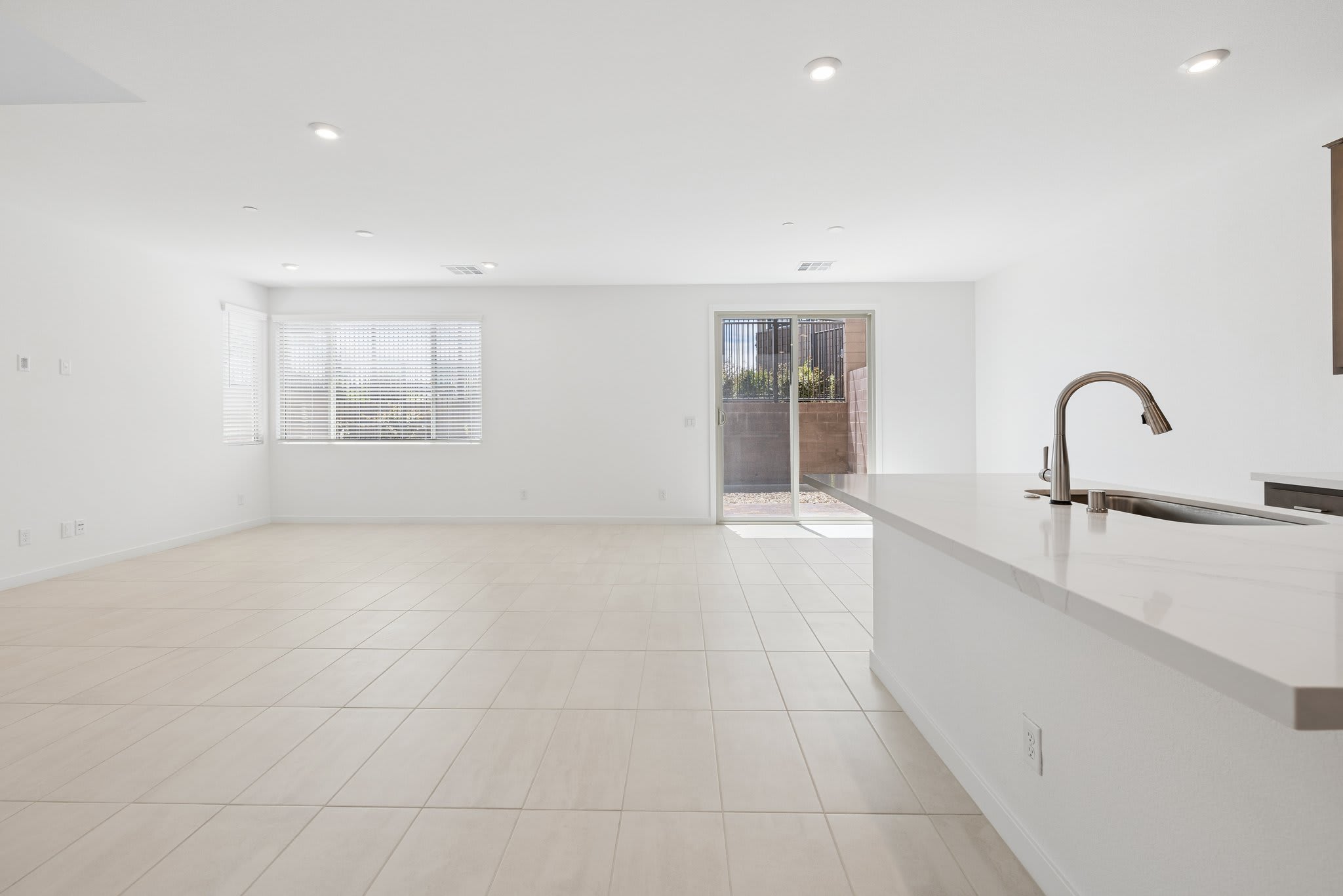 A modern kitchen island with a faucet is in the foreground, while a spacious, brightly lit living area with a sliding glass door leading to a patio is in the background.