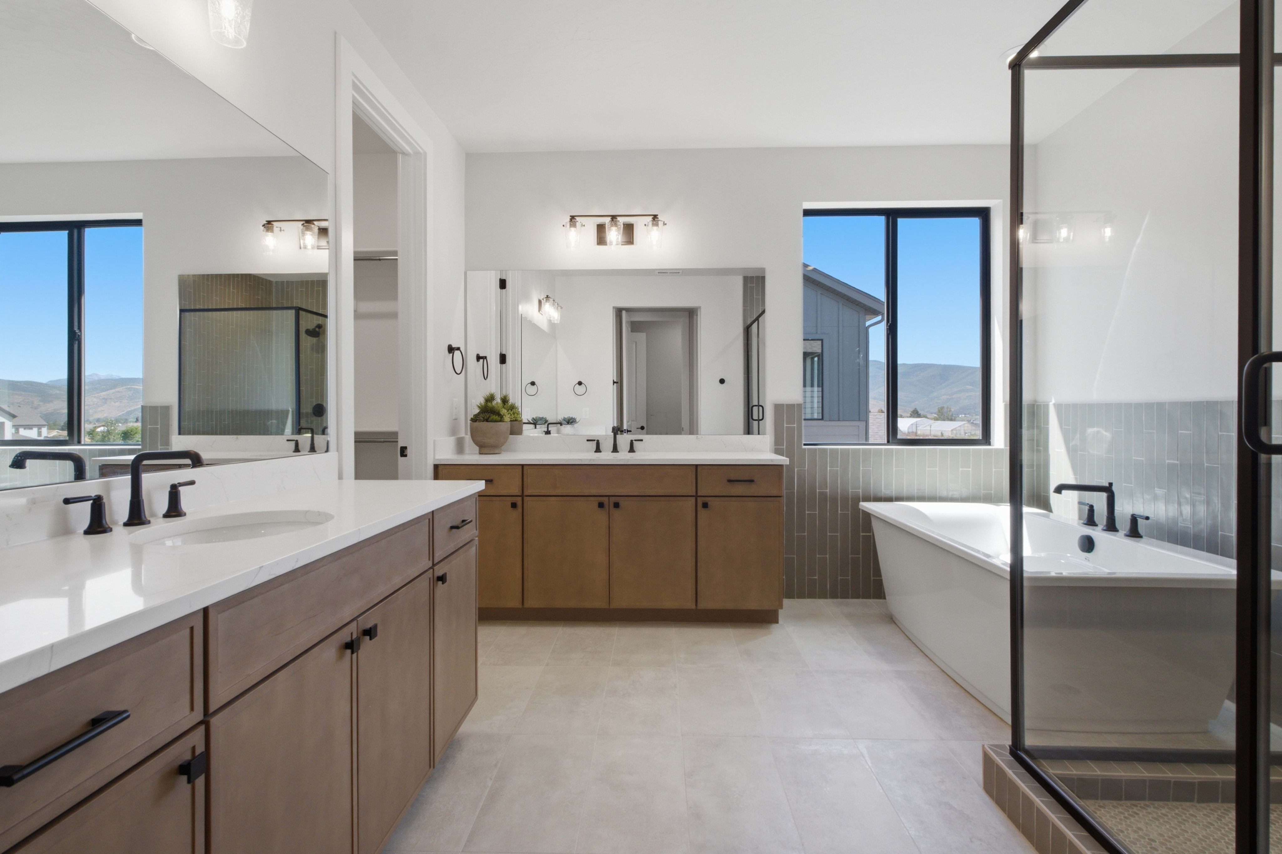 A modern bathroom with a double vanity in the foreground and a freestanding tub and glass-enclosed shower in the background, all illuminated by natural light from large windows.