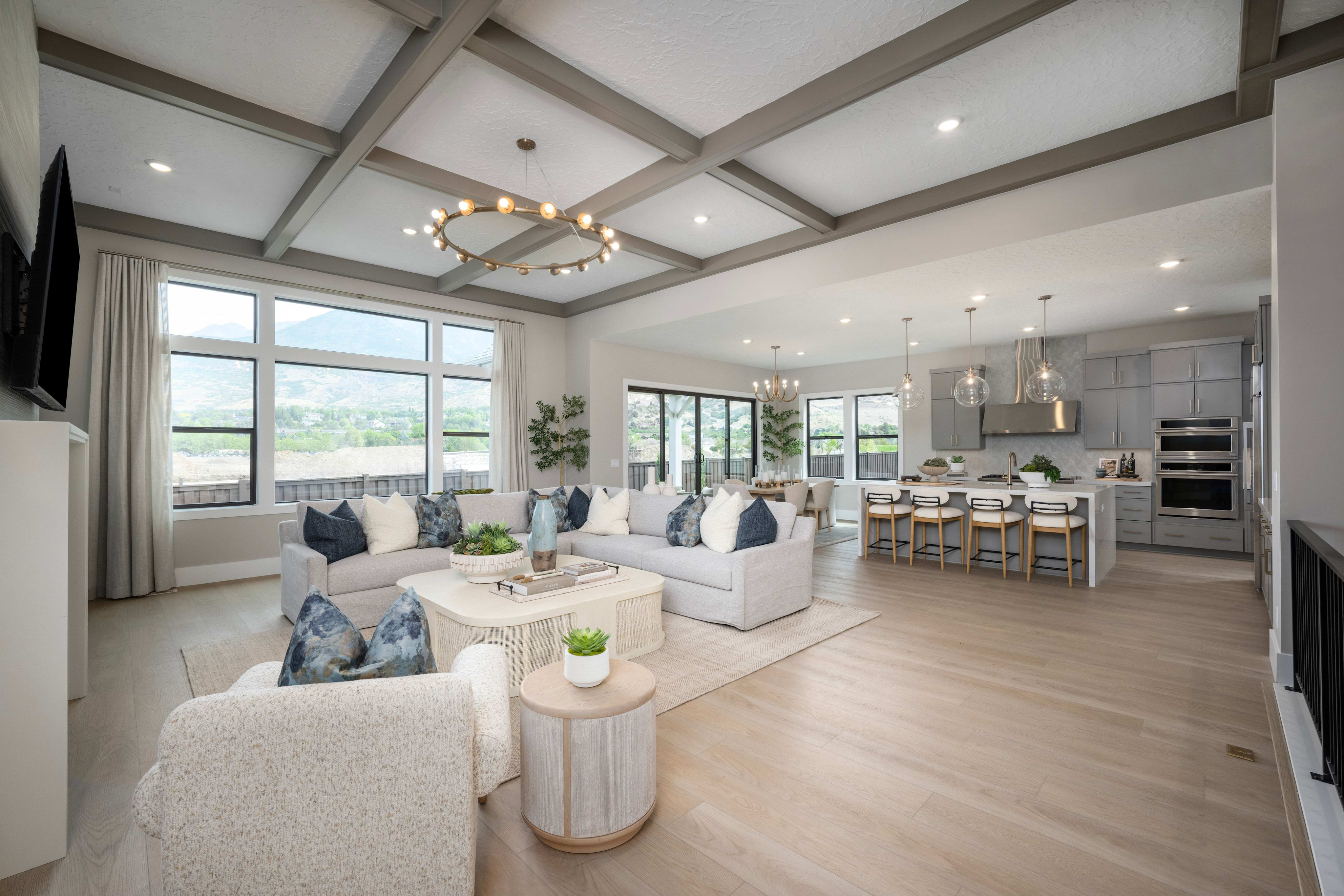A spacious living room with a large sectional sofa and armchairs in the foreground opens to a modern kitchen and dining area in the background, all under a coffered ceiling with a chandelier.