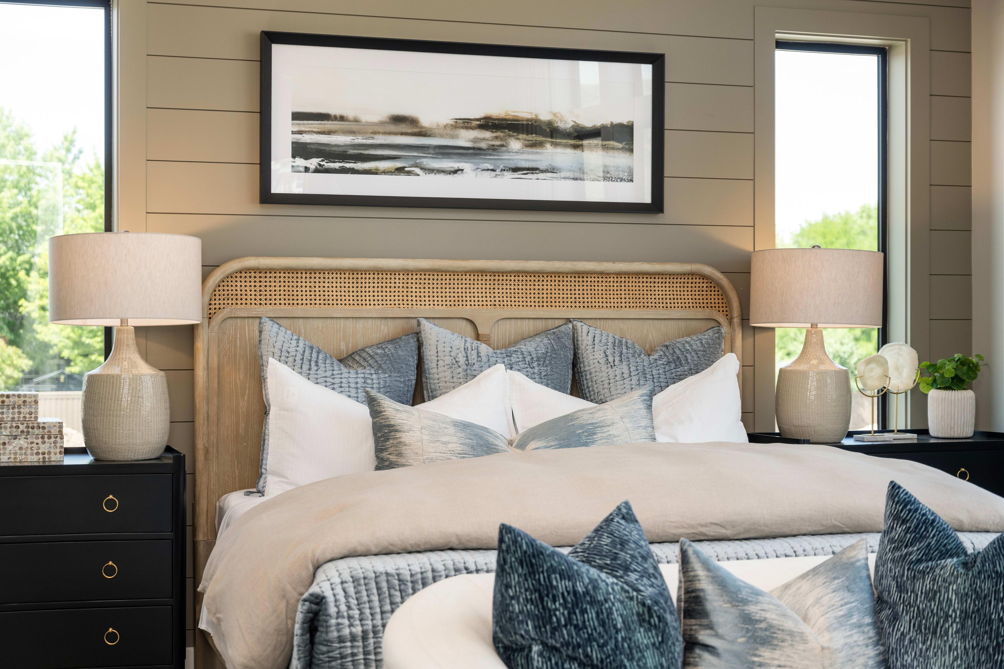 A cozy bedroom features a plush bed with blue and white pillows in the foreground, while a large abstract landscape painting hangs above the headboard against a backdrop of neutral-toned walls and windows.