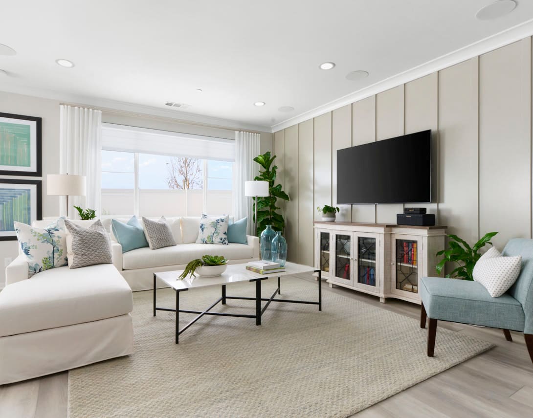 A modern living room features a white sectional sofa and a coffee table in the foreground, with a television and a light blue armchair against a paneled wall in the background.