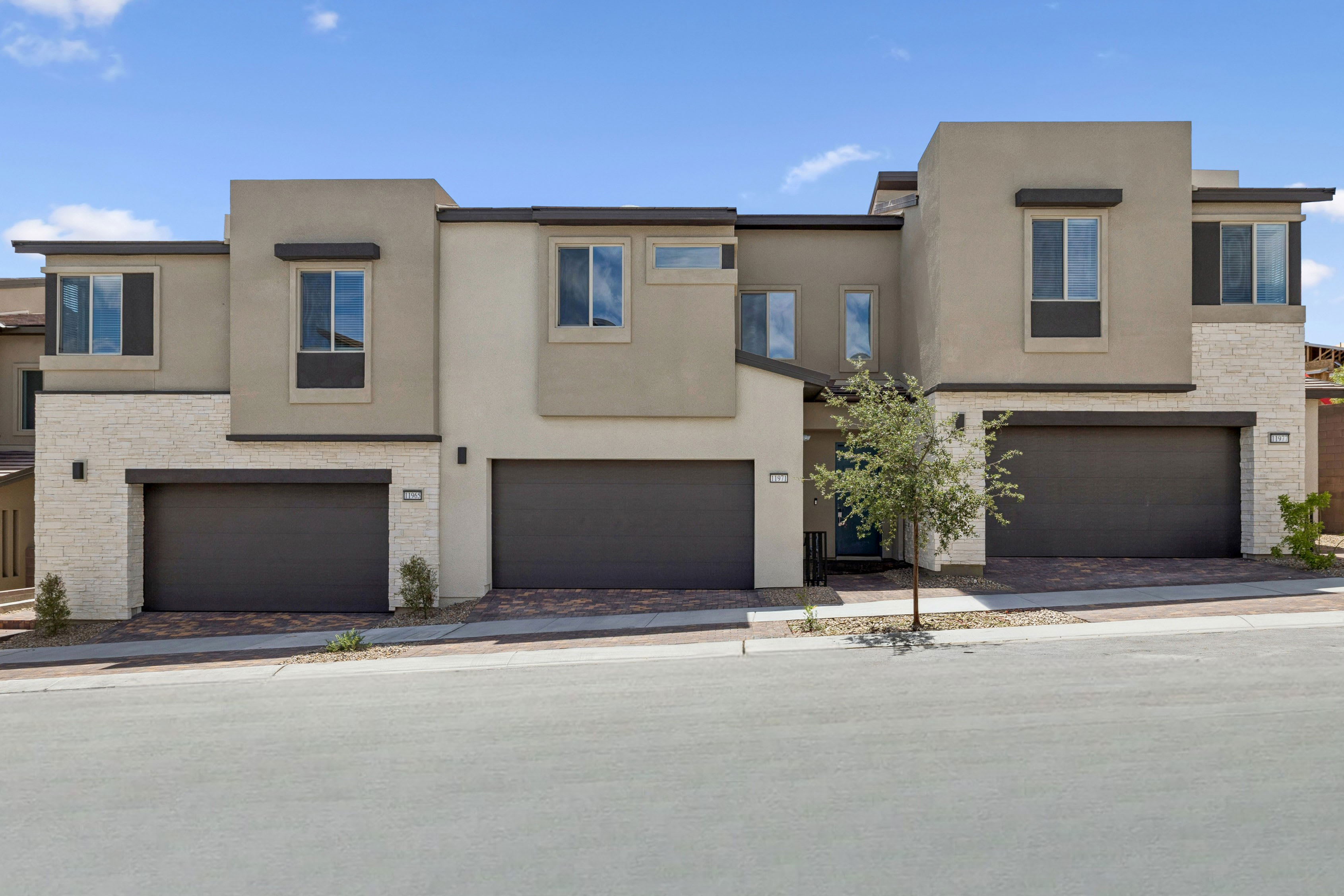 A modern townhouse complex with three garages in the foreground sits against a backdrop of a clear blue sky with scattered clouds.