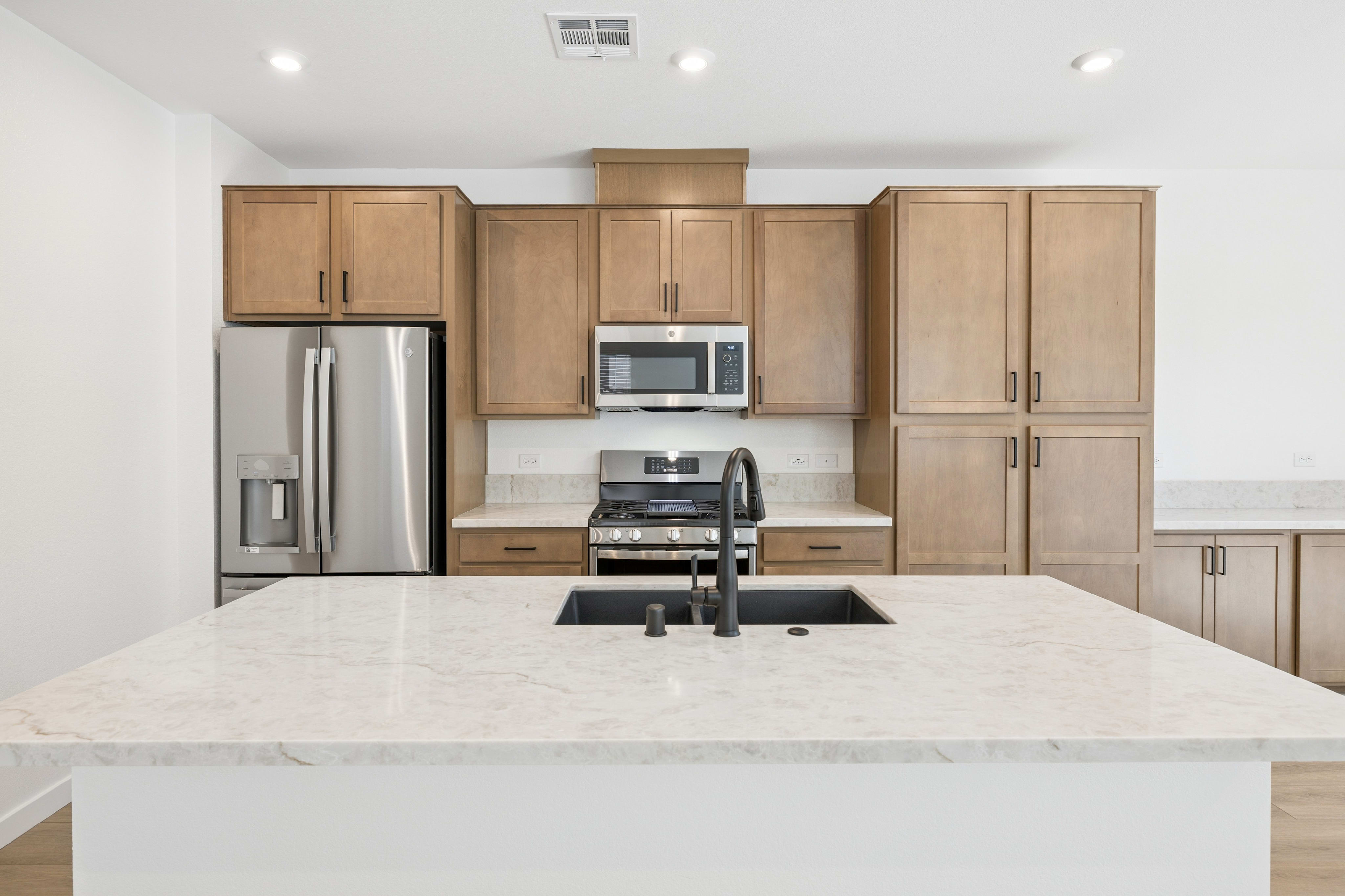 A modern kitchen island with a marble countertop is in the foreground, while stainless steel appliances and light wood cabinets fill the background.