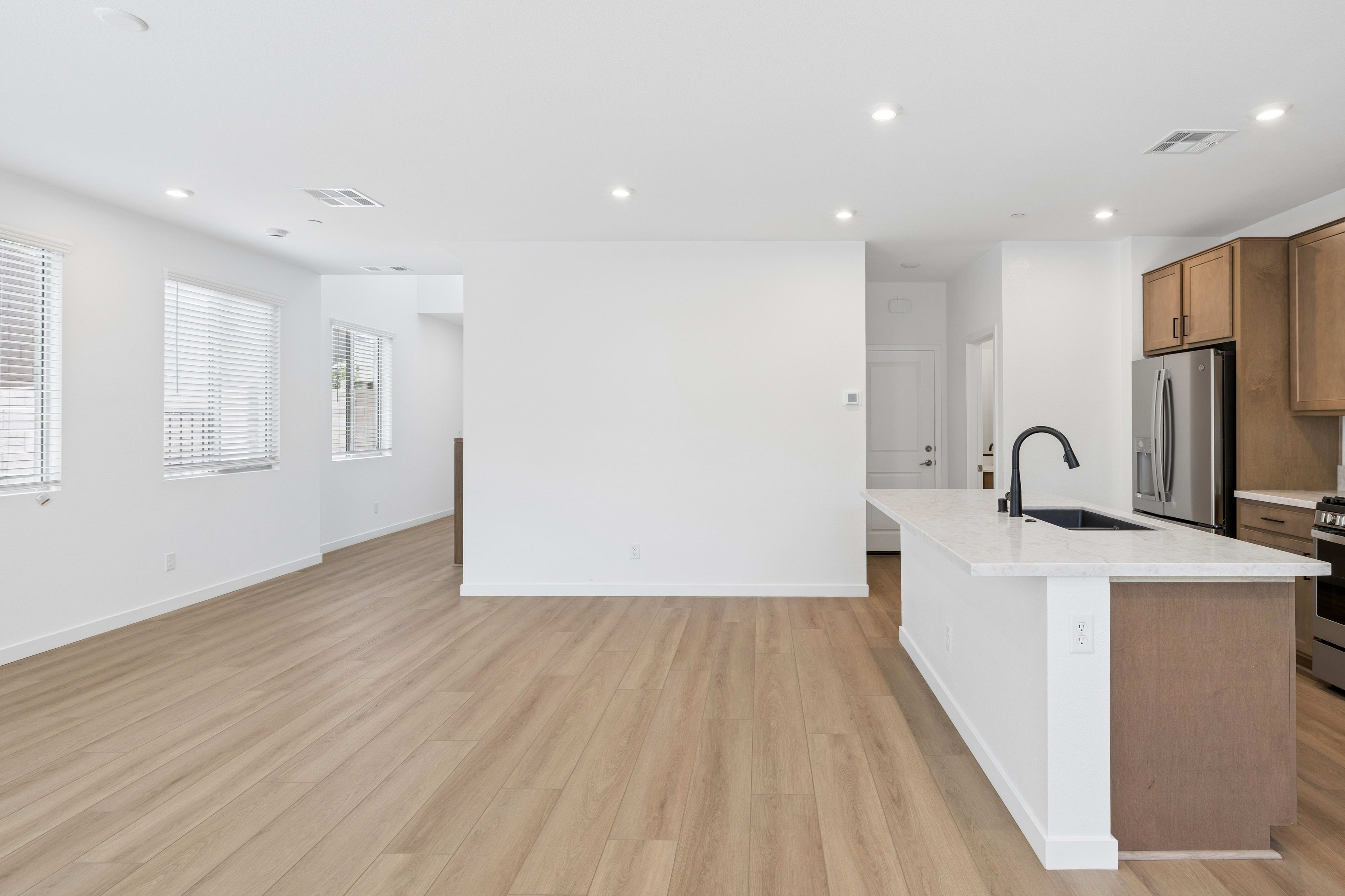 A modern kitchen island with a black faucet stands in the foreground, while a spacious, brightly lit living area with large windows and light wood flooring extends into the background.