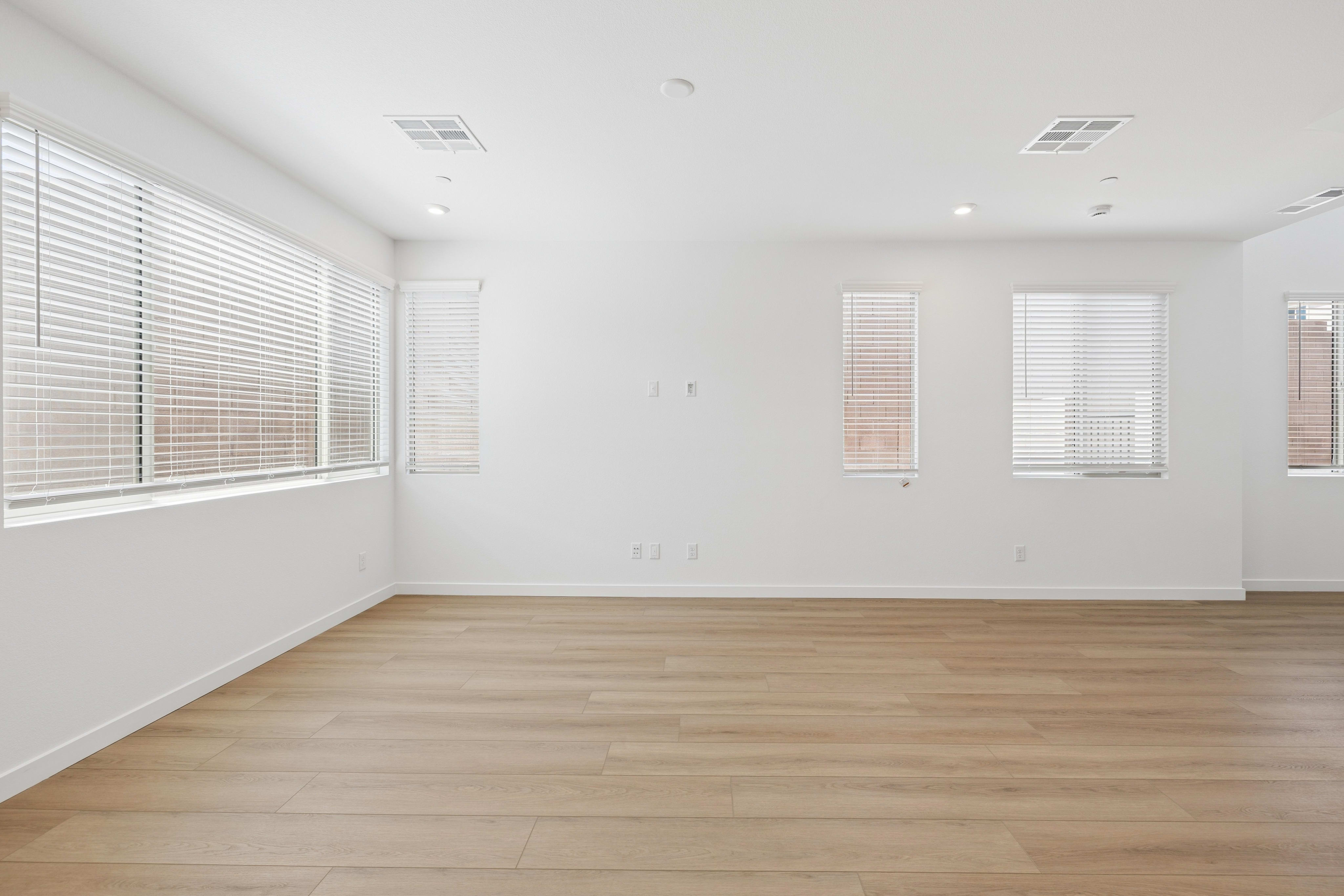 A bright, empty room with light wood flooring in the foreground and white walls with blinds-covered windows in the background.