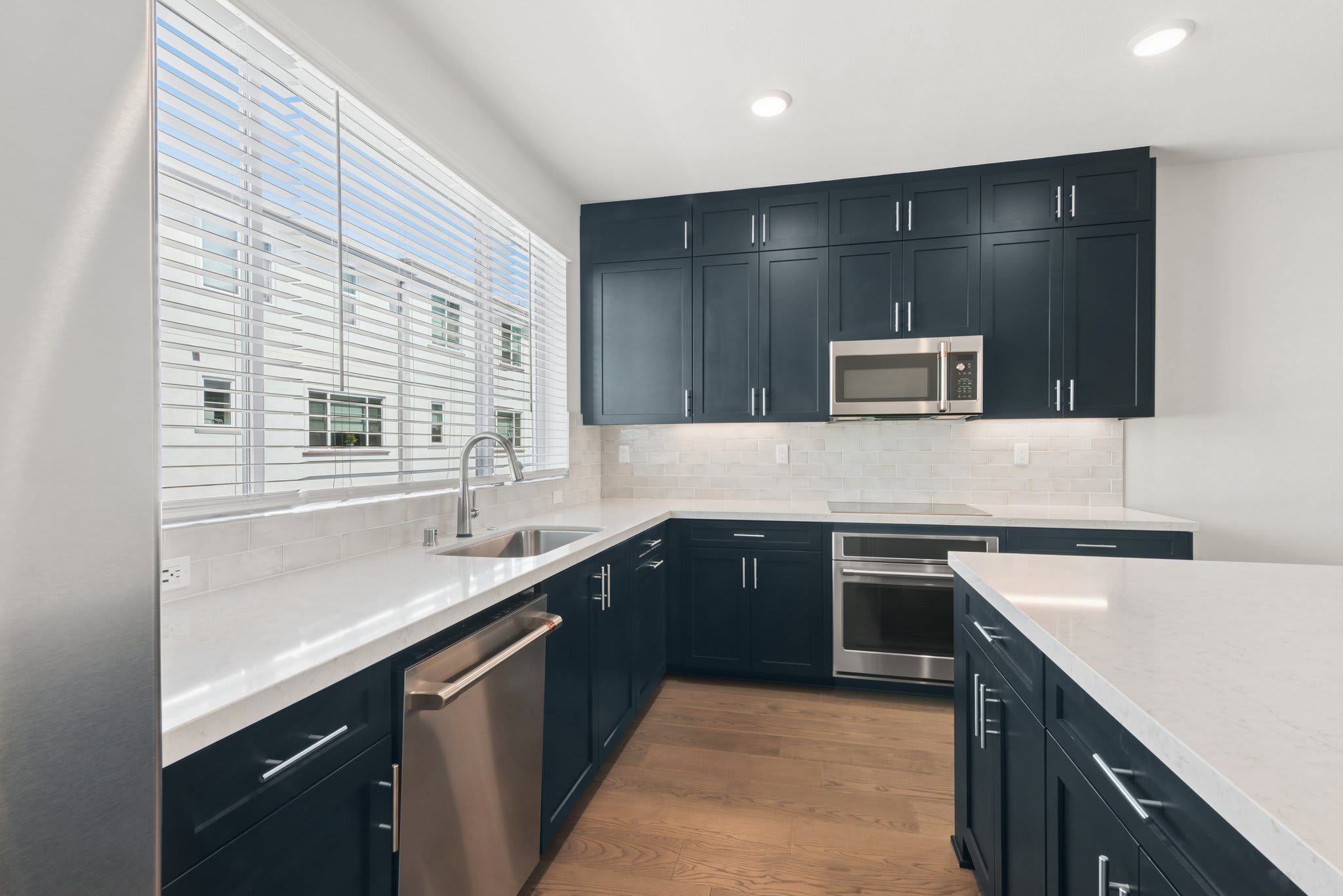 A modern kitchen with dark blue cabinets and white countertops features a large window with blinds in the foreground and a stainless steel oven and microwave in the background.