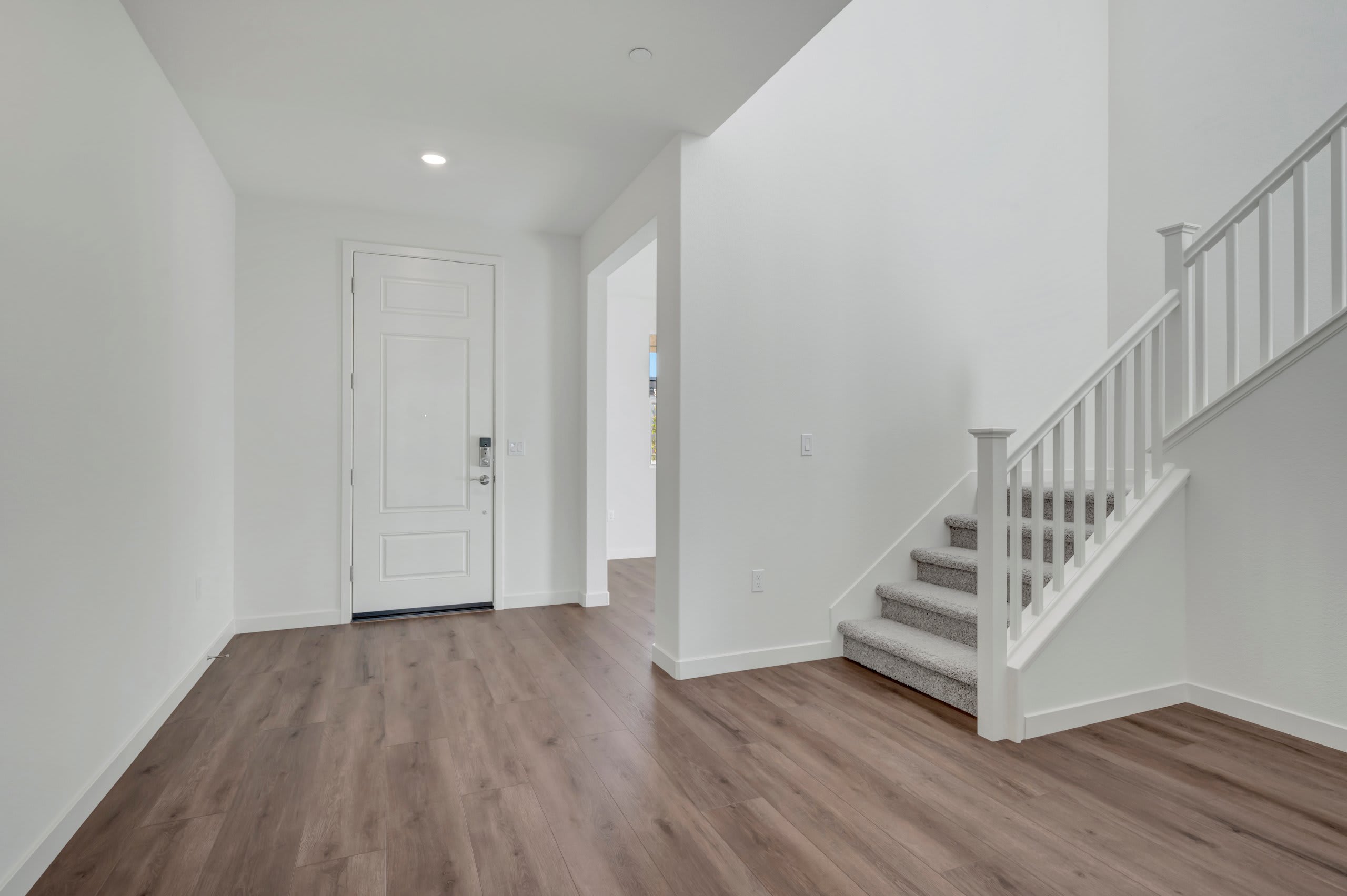 A white front door stands in the foreground with a gray carpeted staircase to the right, leading up to a bright, empty hallway in the background.