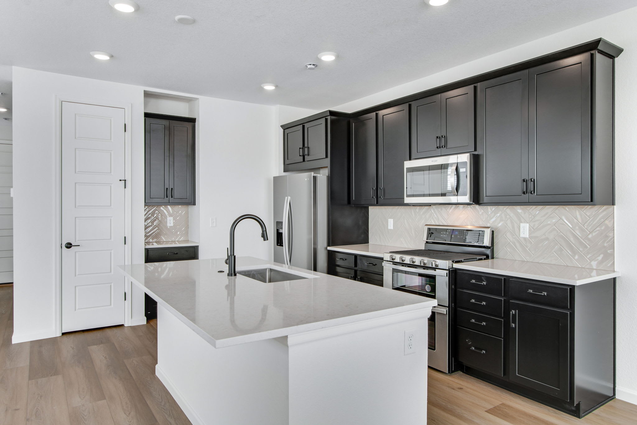 A modern kitchen with a white island in the foreground and dark cabinetry and stainless steel appliances in the background.