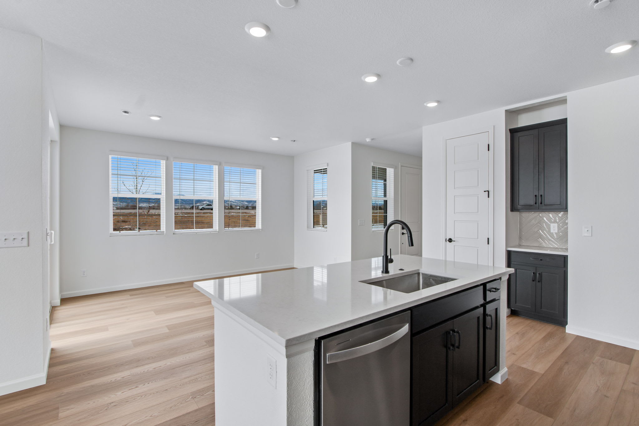 A modern kitchen island with a sink and dishwasher is in the foreground, while a living area with large windows overlooking a landscape is in the background.