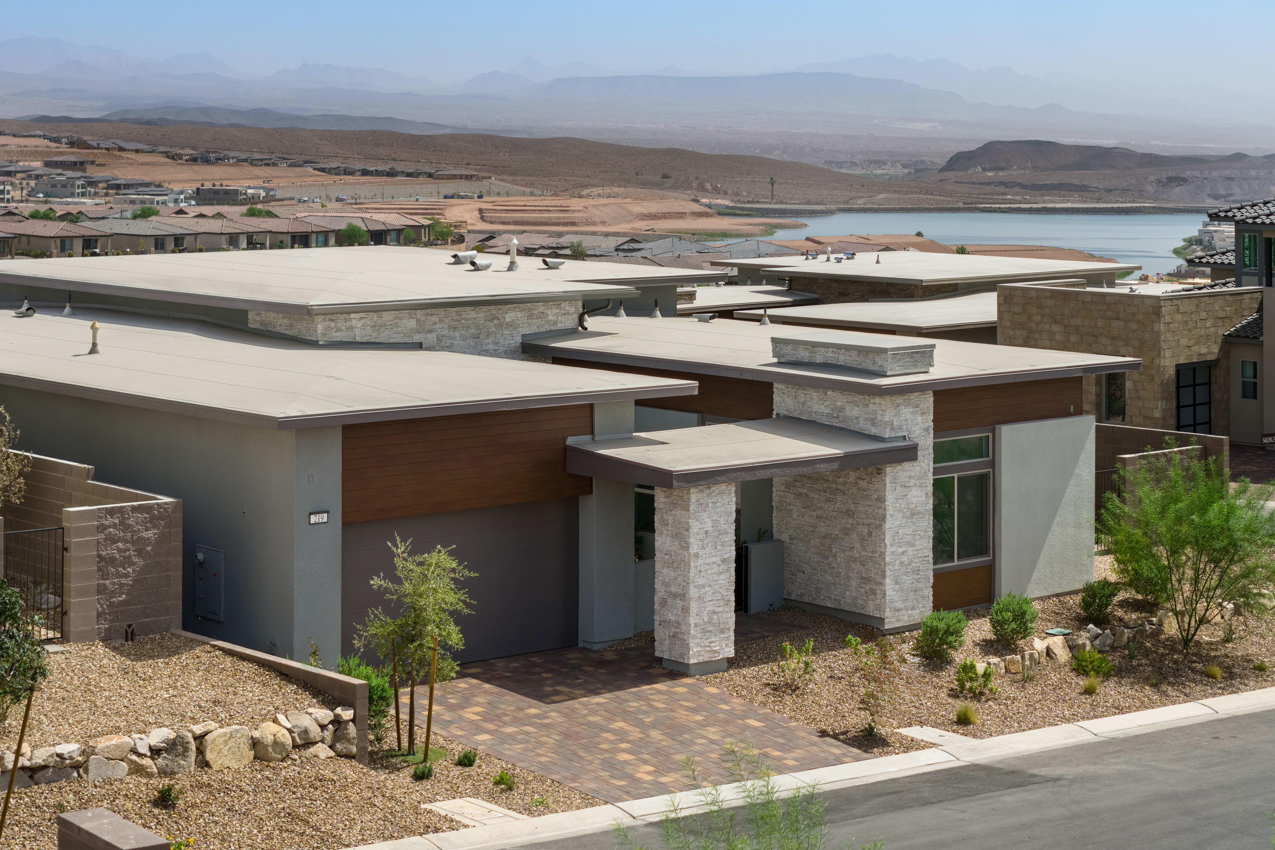 Modern homes with flat roofs and stone accents are situated in a dry, arid landscape with a distant lake and mountains.