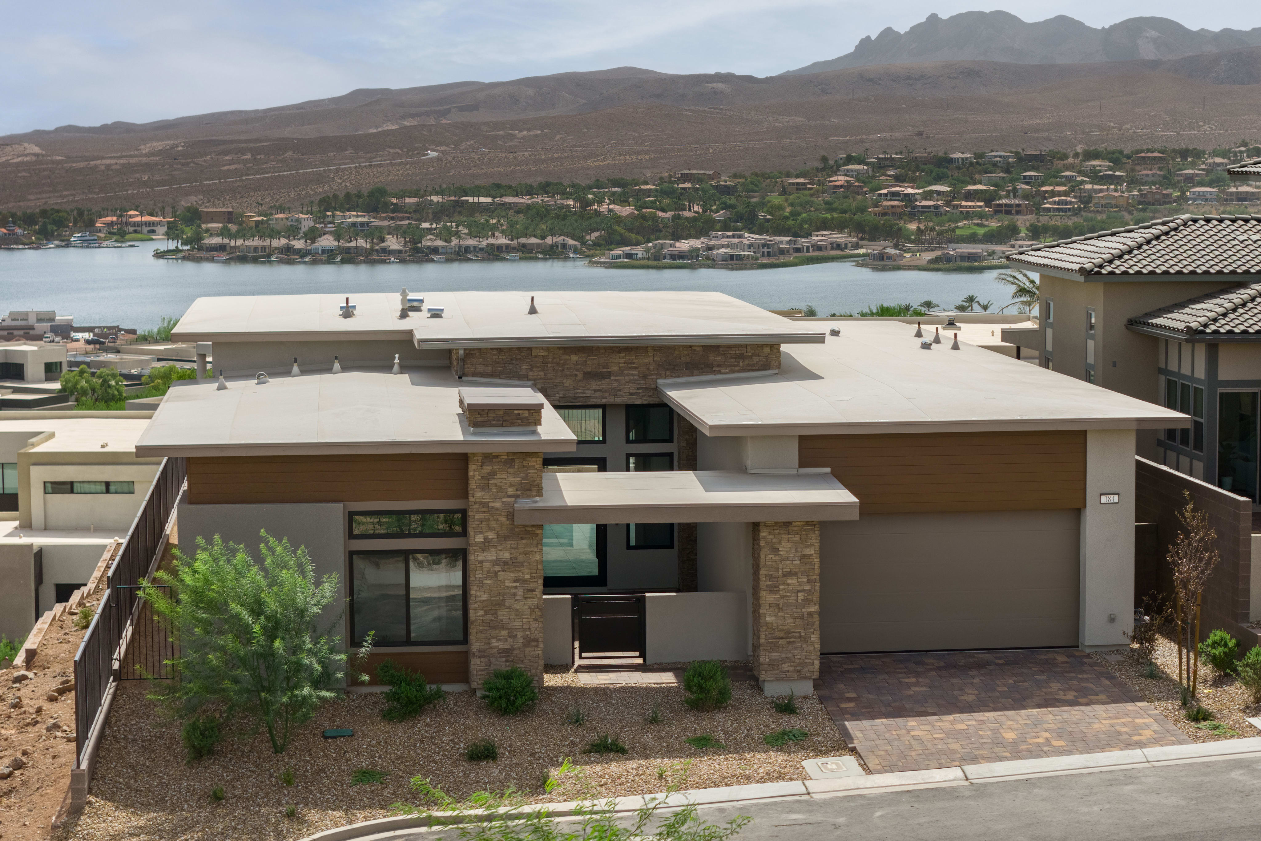 A modern house with stone accents sits in the foreground, overlooking a tranquil lake and distant, arid mountains.