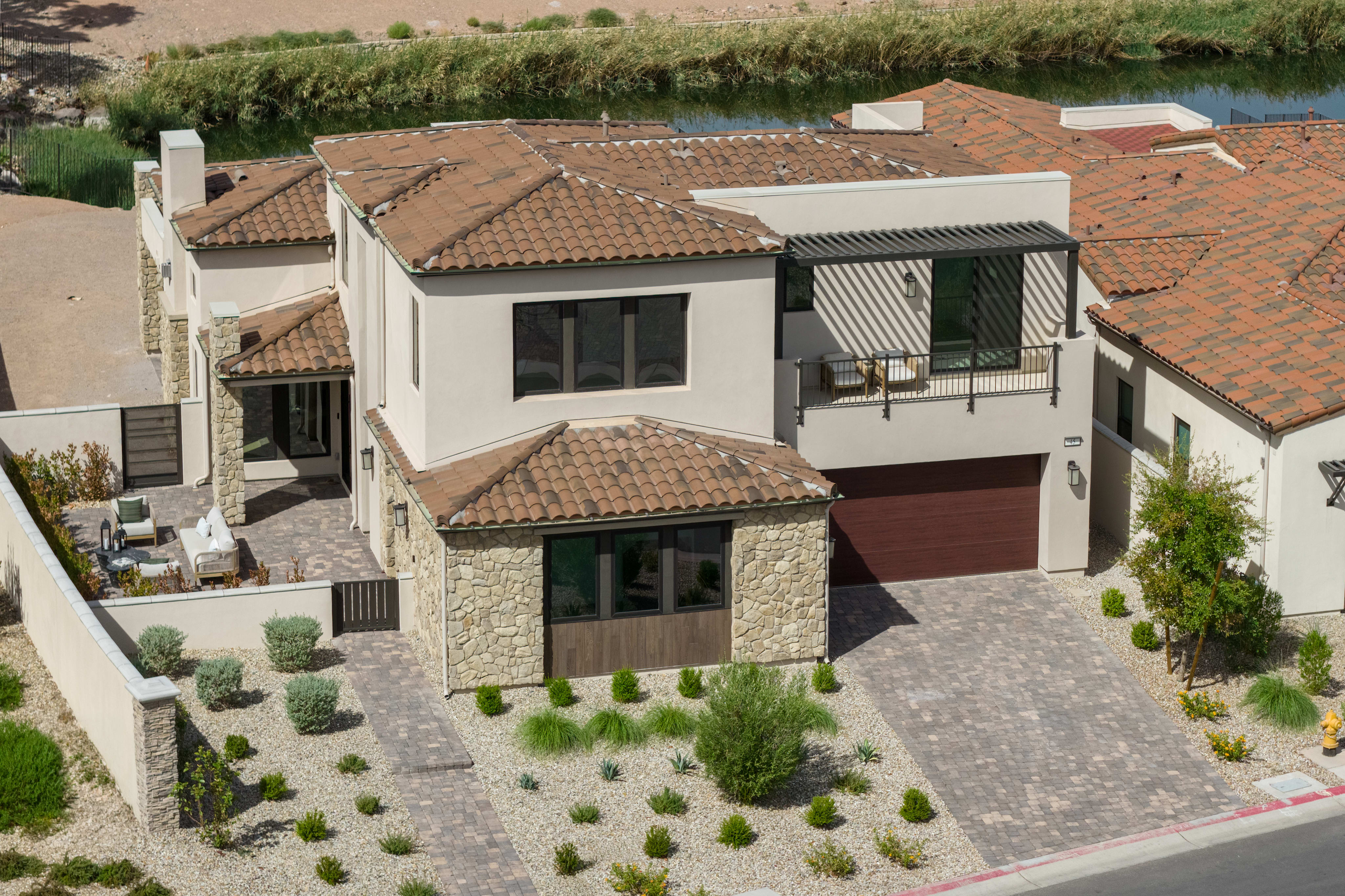 A modern stucco and stone house with a tiled roof sits in the foreground, with a calm body of water and reeds visible in the background.
