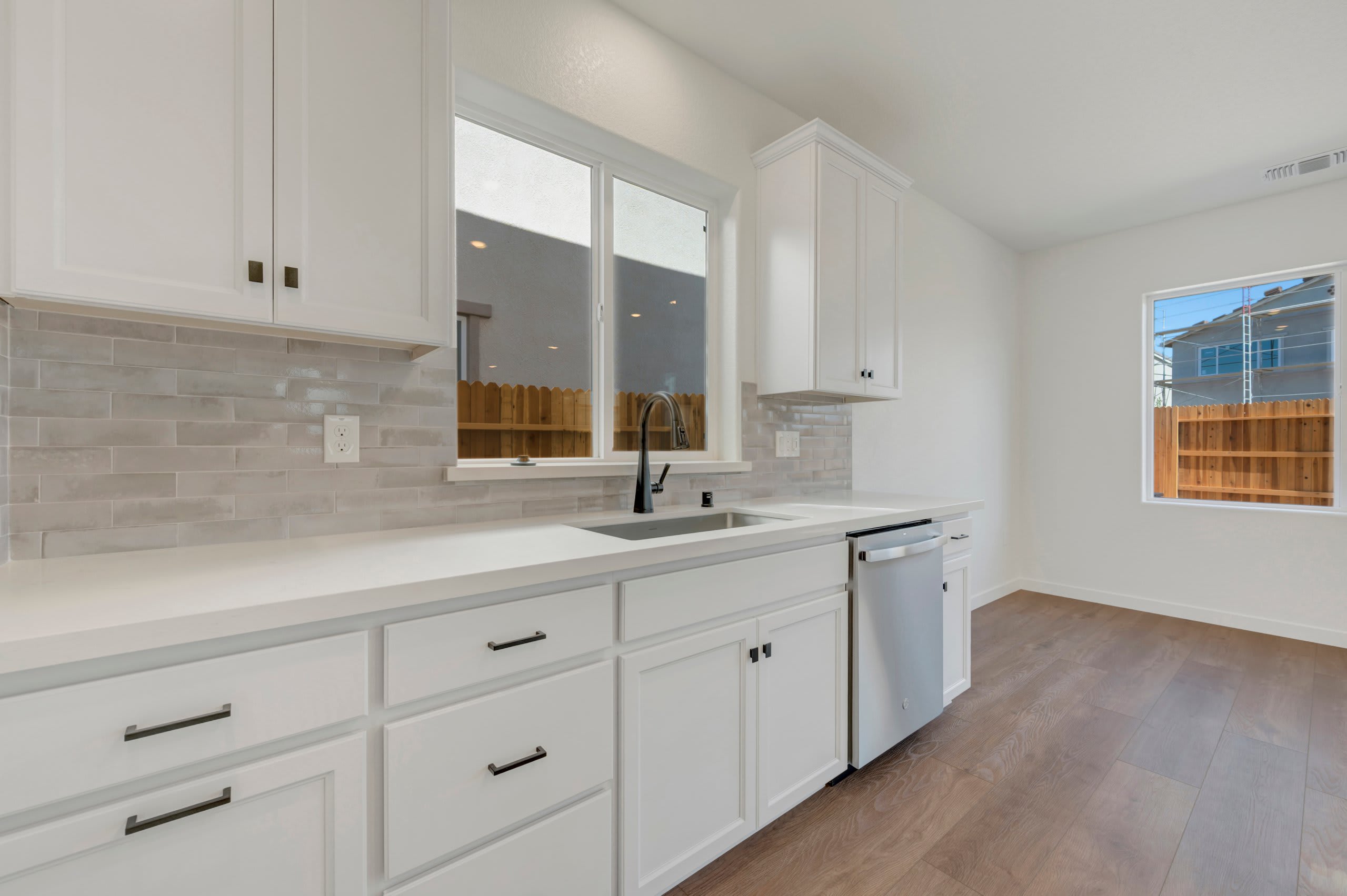 White cabinets and a tiled backsplash frame a sink and dishwasher in the foreground, while a wooden floor leads to a window with a view of houses and a fence in the background.